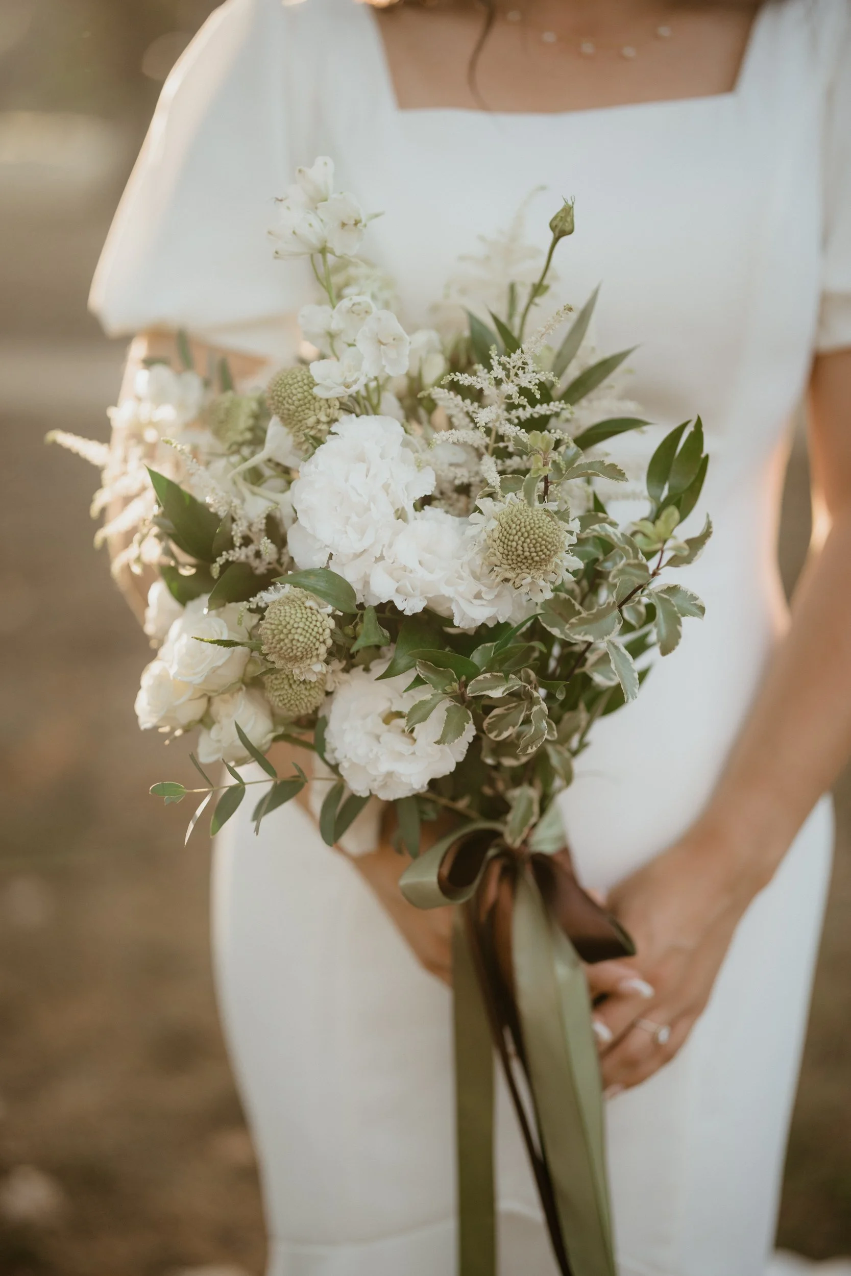 A person in a white dress holding a bouquet of white and cream flowers with greenery, tied with a brown ribbon.