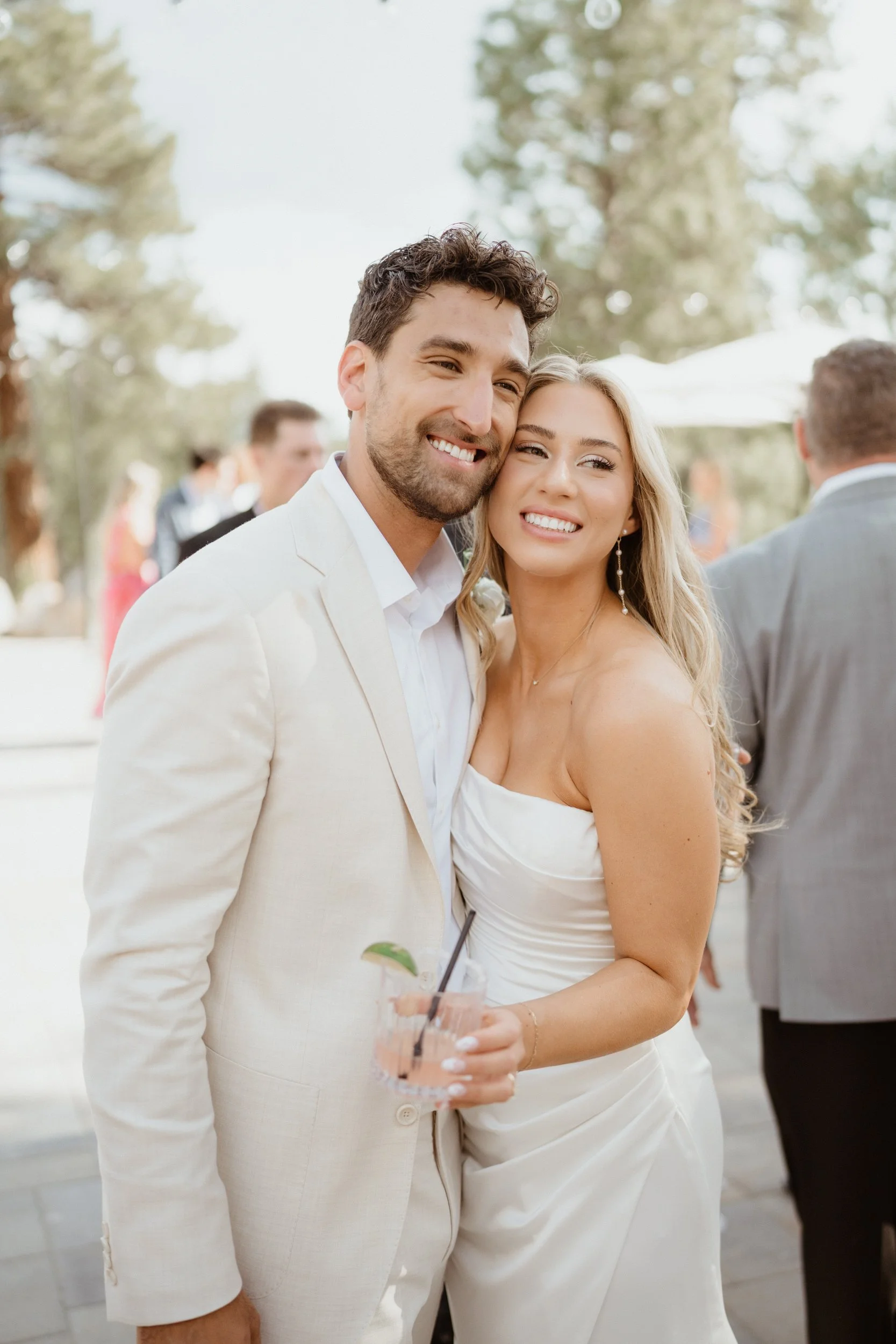 A smiling couple dressed in wedding attire at an outdoor event, with trees in the background. The man is wearing a cream suit, and the woman is in a white strapless dress holding a drink with a lime wedge.