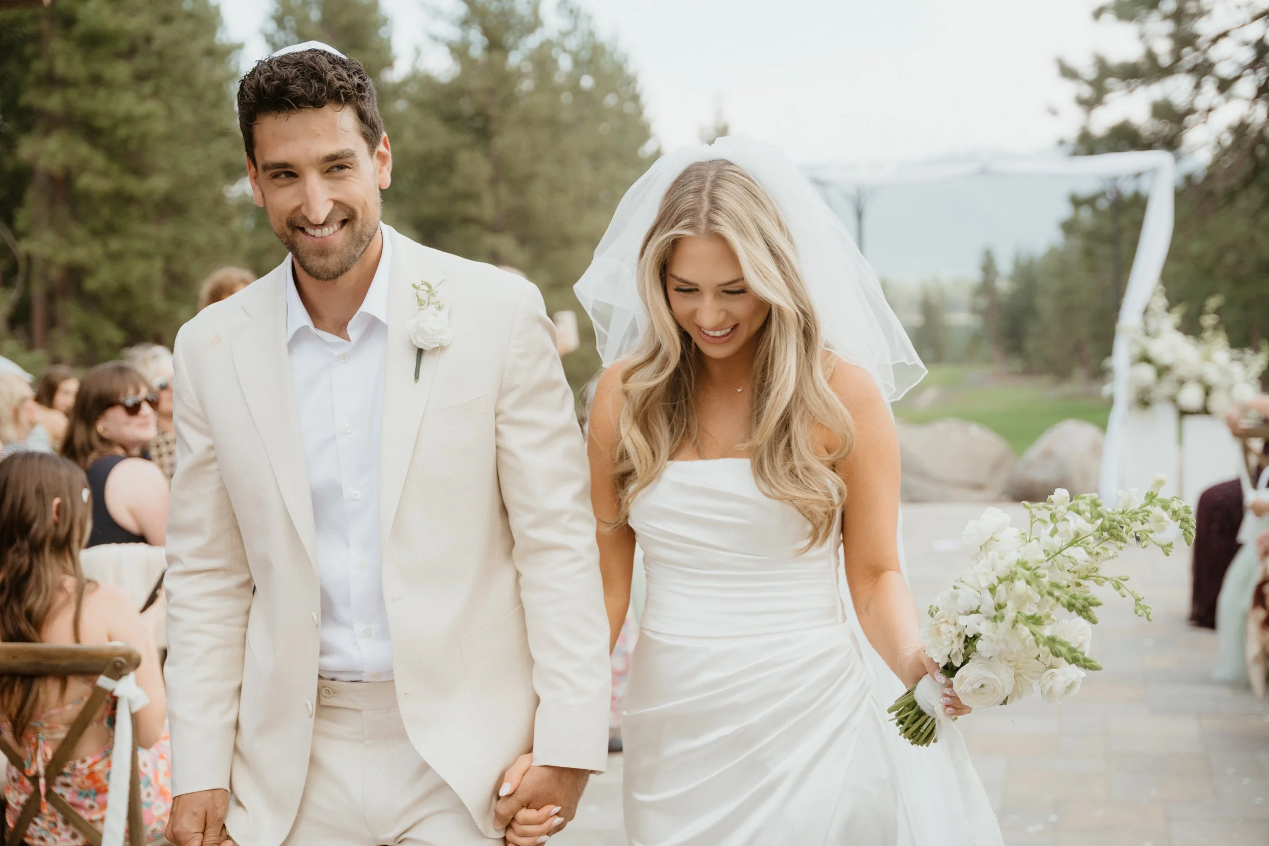 A bride and groom holding hands and smiling during their wedding ceremony outdoors.