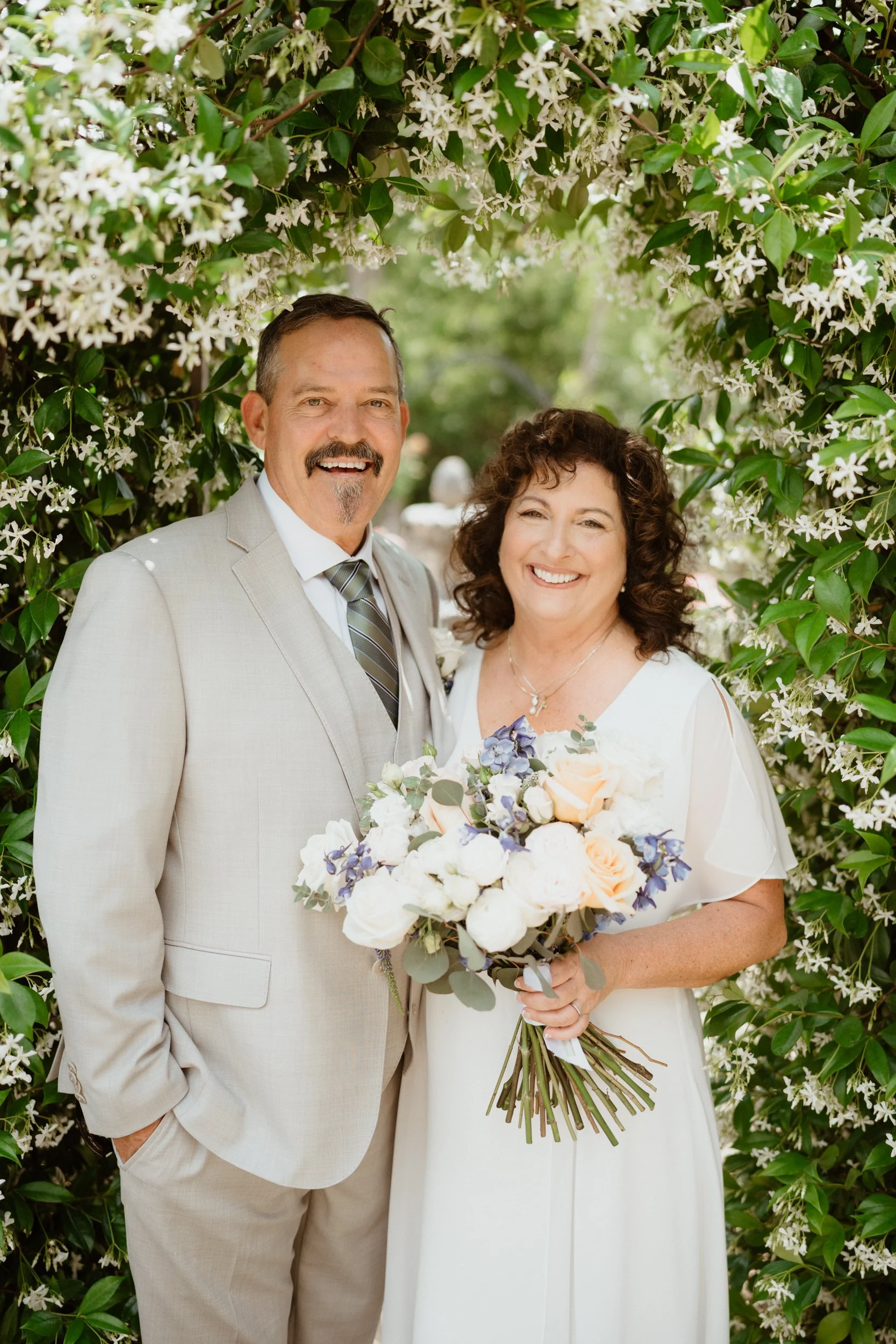 A smiling couple in wedding attire standing under a floral archway outdoors, holding a bouquet of flowers.