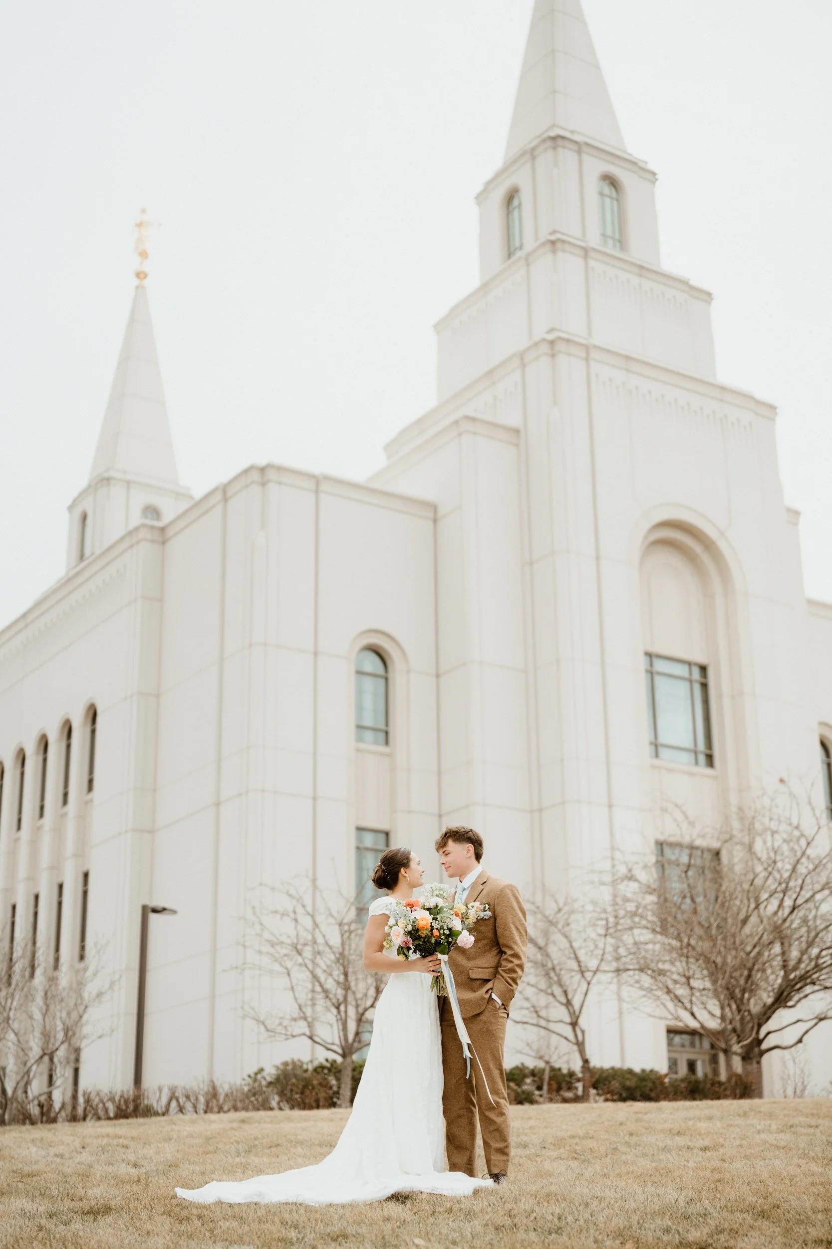 A bride and groom standing on grass in front of a white church, smiling and holding a bouquet of flowers.