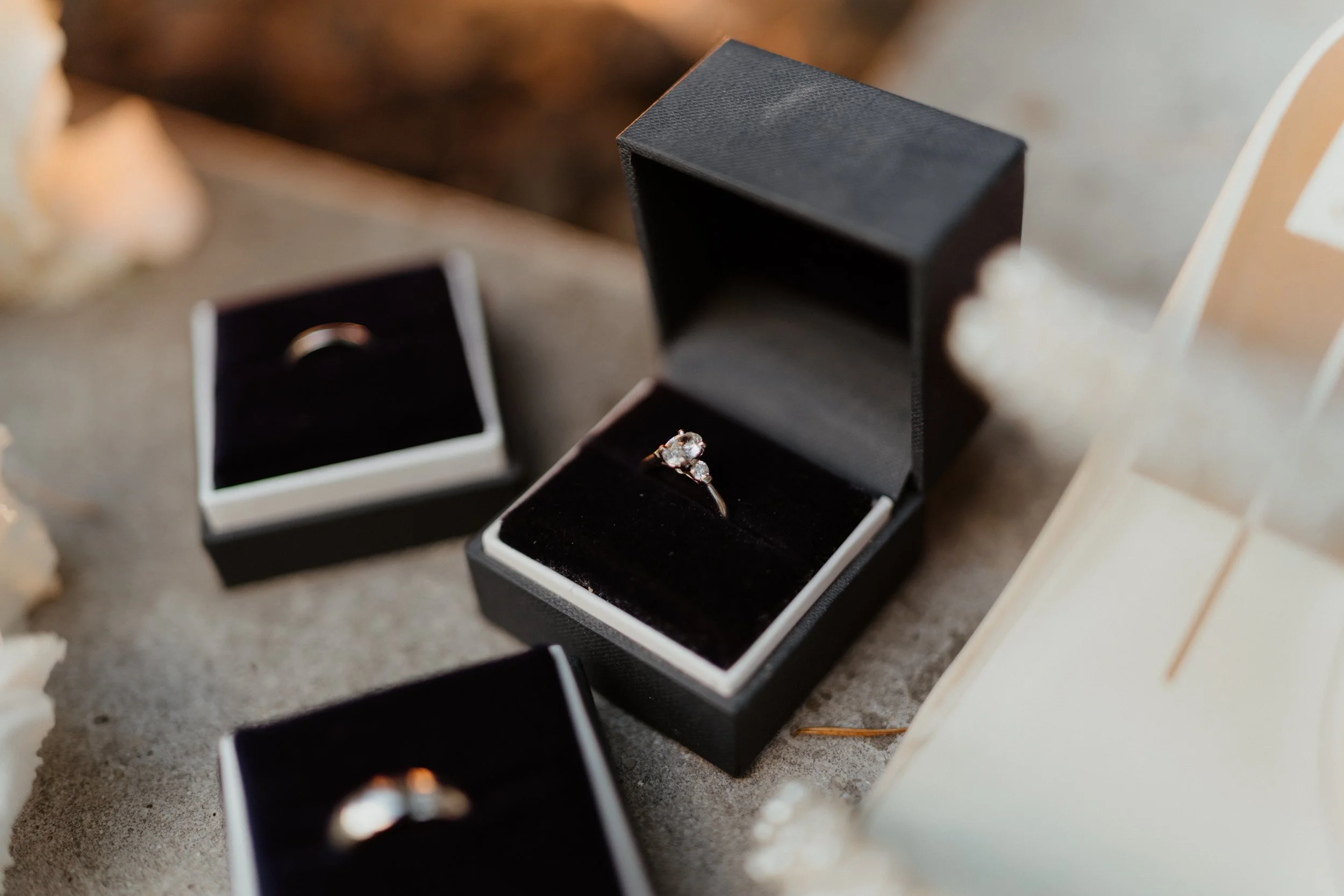Close-up of a diamond engagement ring in a black jewelry box, surrounded by other jewelry boxes and a white photo frame on a textured surface.