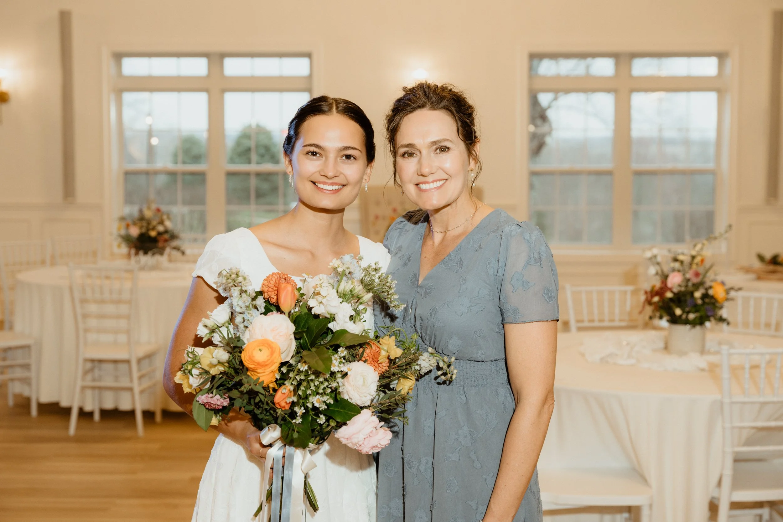 A woman in a white dress holding a bouquet of flowers standing next to another woman in a blue dress inside a decorated event room with large windows.
