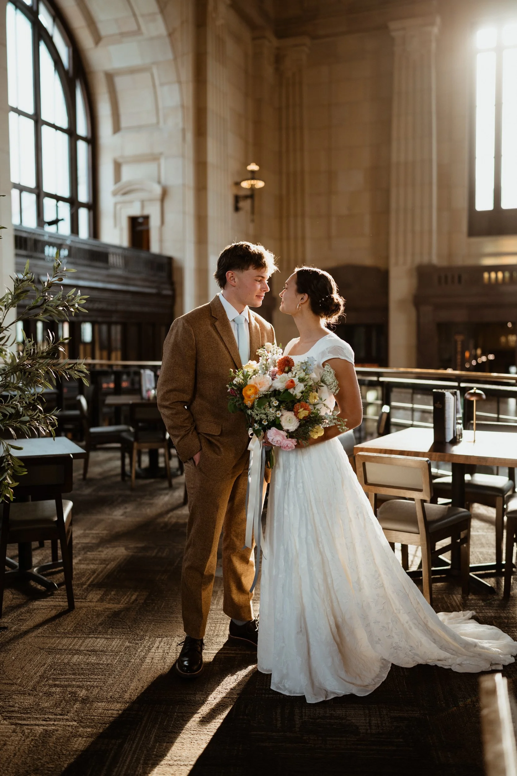 A bride and groom standing close together in a spacious, sunlit room with tall windows and classical architecture, holding a bouquet of flowers.