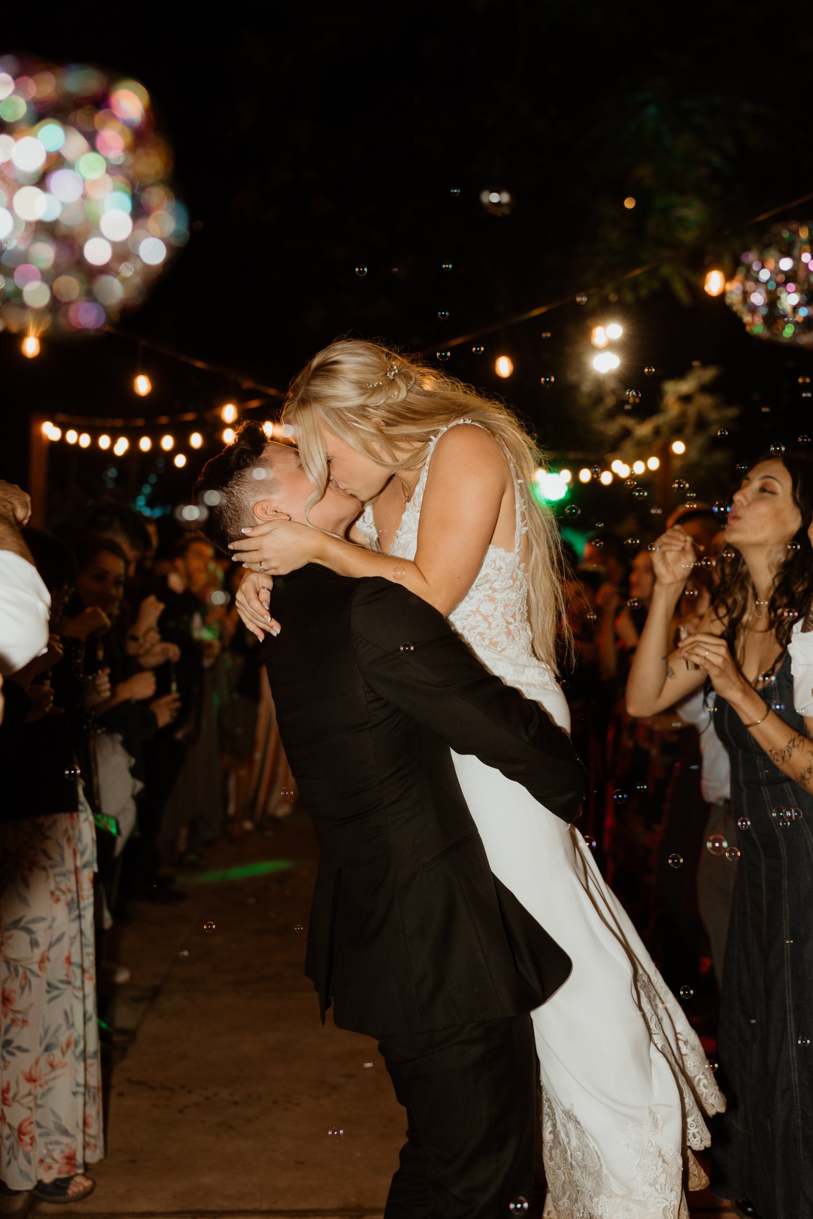 A couple kissing at a nighttime celebration, possibly a wedding, with guests around and string lights overhead.