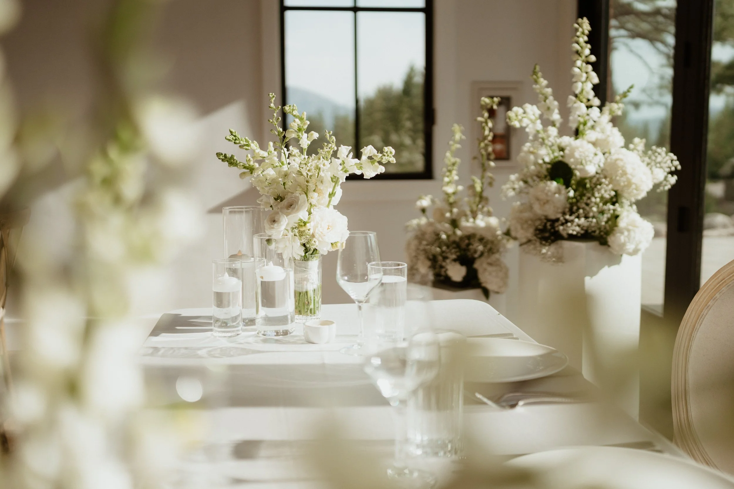 Elegant dining table set with white floral arrangements, glassware, and a white tablecloth, with large windows and scenic outdoor view in the background.