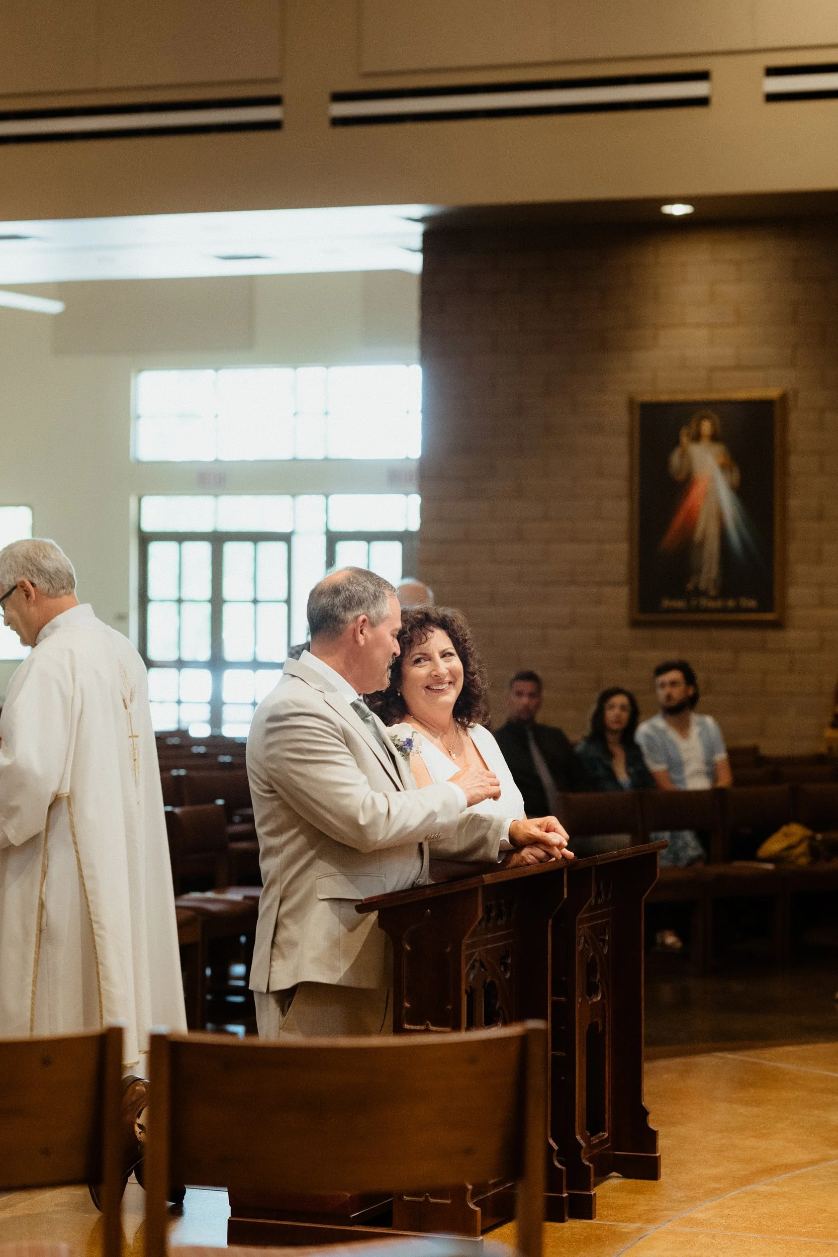 A couple stands at a church altar during a wedding ceremony, smiling and looking at each other, with a priest on the left and bridesmaids and groomsmen seated in the background.