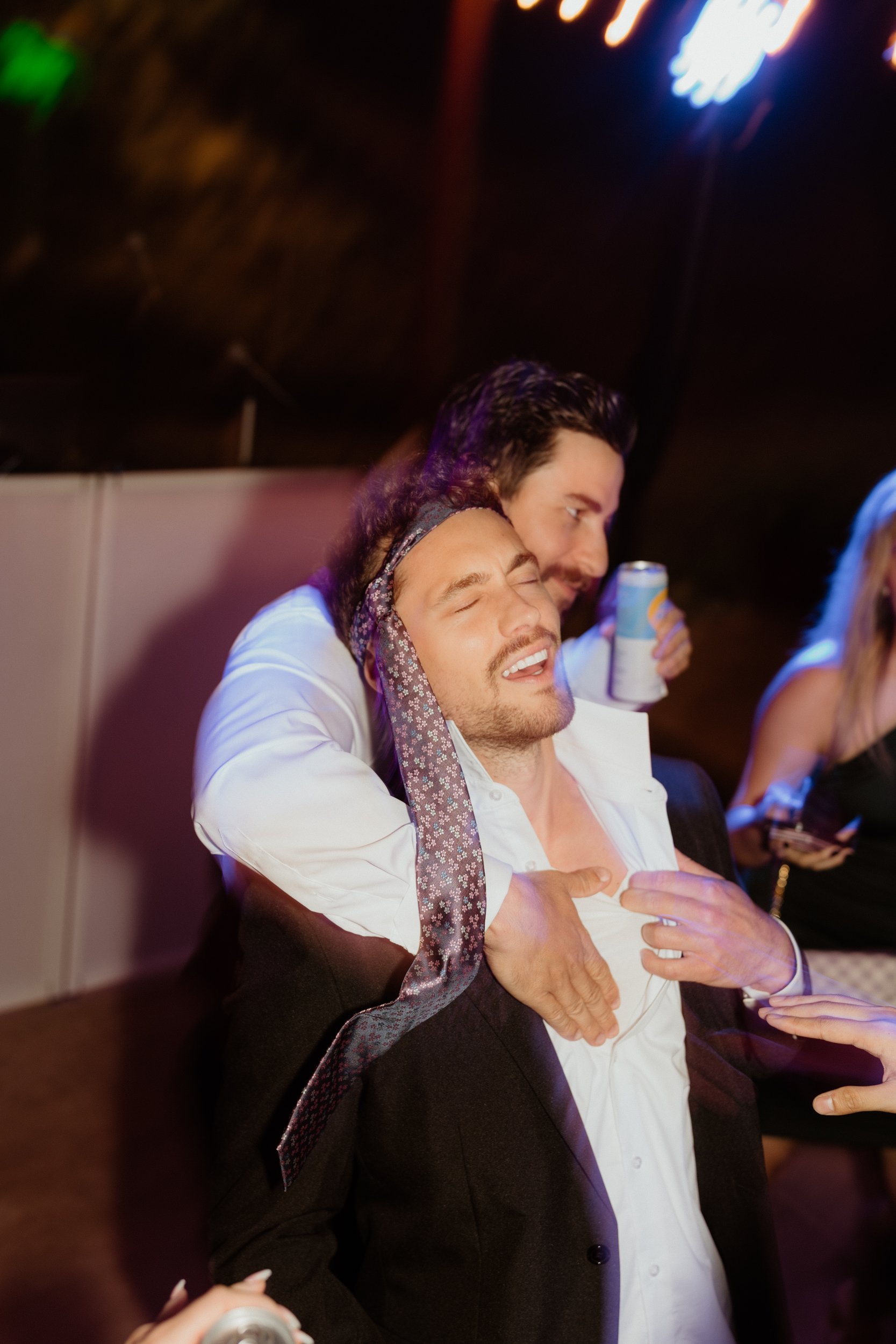 Two men, one with long dark hair and the other with short brown hair, are in a playful pose; the man with long hair appears to be choking or pretending to choke the other man, who has his eyes closed and a pained expression, while holding a can of be