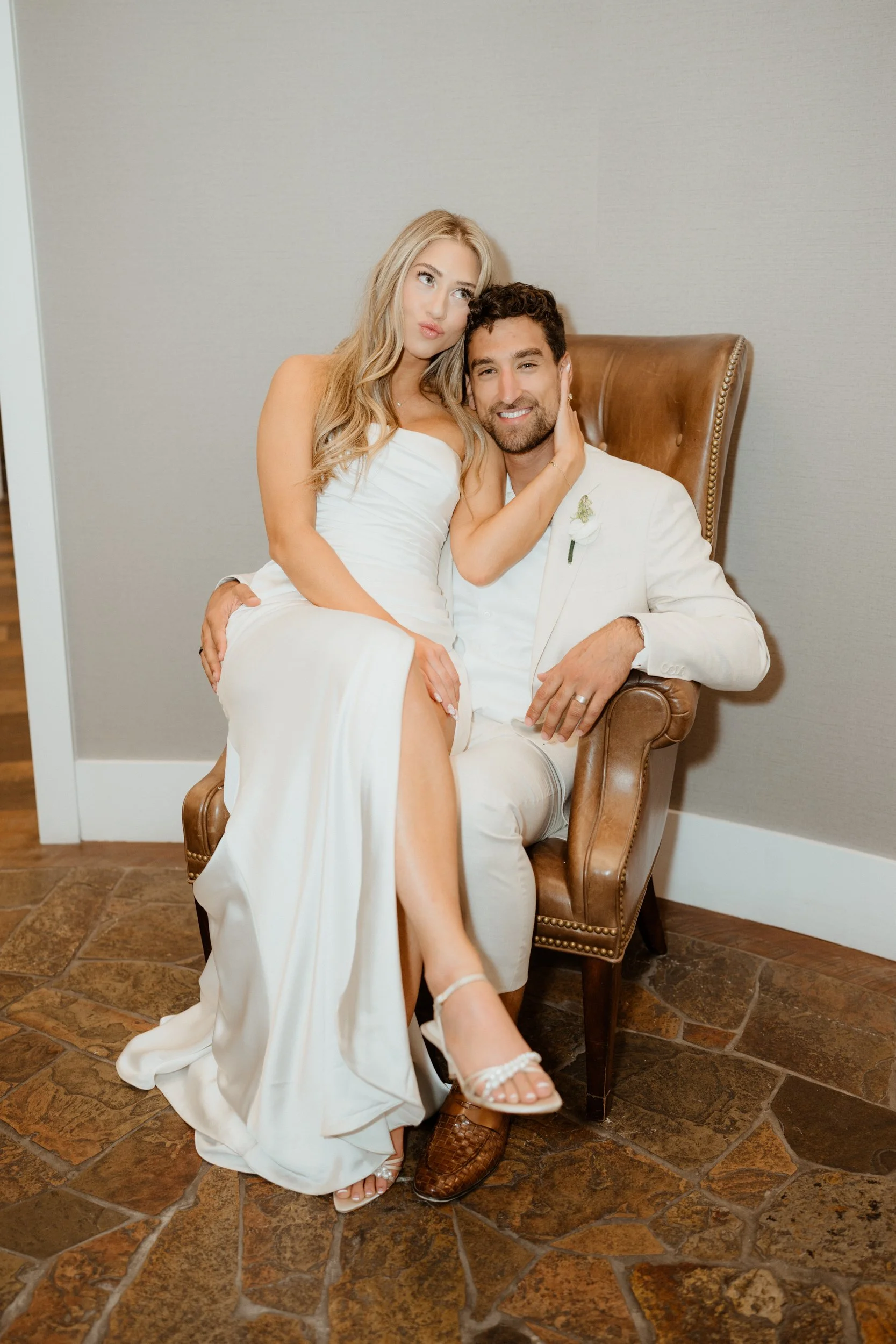A bride and groom sitting together in a wedding photo, with the bride wearing a white dress and the groom in a white suit, in an indoor setting.