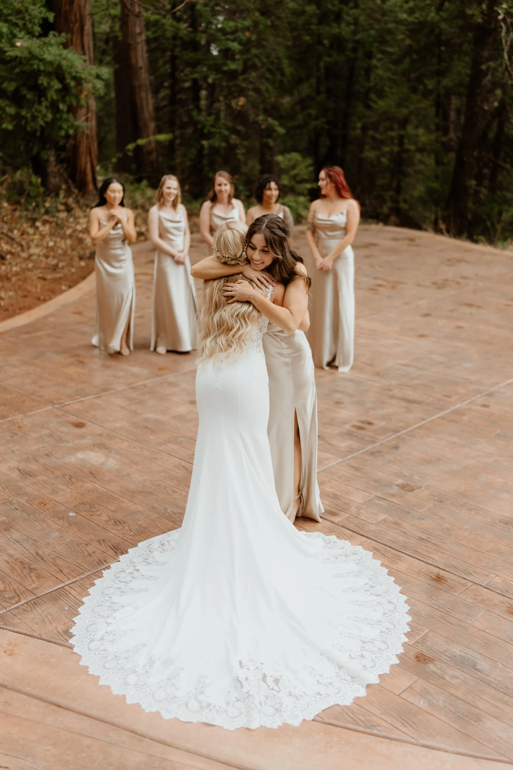 A bride with long, dark hair hugging her mother, both smiling, on an outdoor wooden dance floor surrounded by trees. Four bridesmaids in matching beige dresses watch and smile in the background.