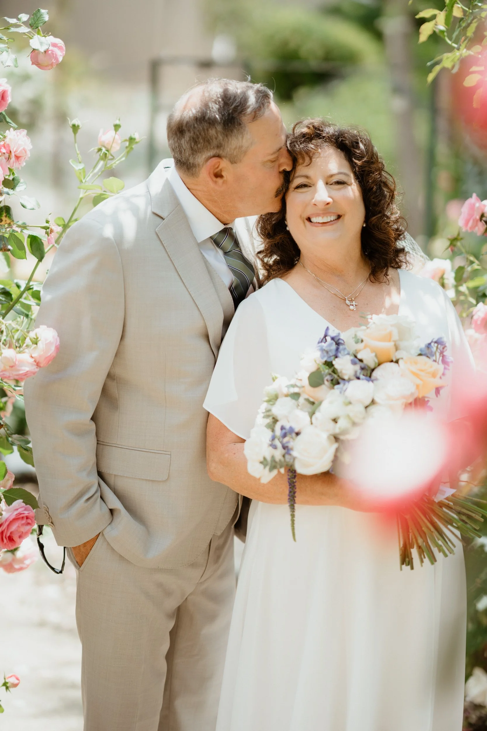 A man in a beige suit kisses a smiling woman in a white dress holding a large bouquet of flowers, standing among pink and white roses on a sunny day.
