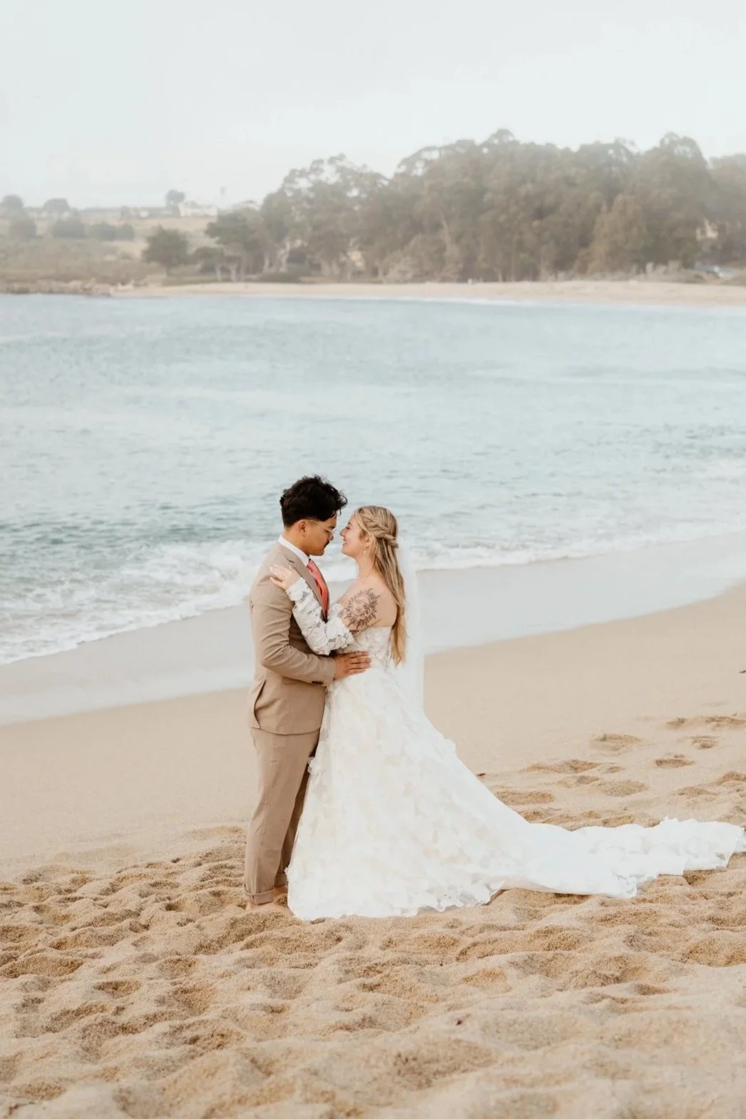 A bride and groom standing closely together on a beach, with the groom in a beige suit and the bride in a white wedding gown, touching foreheads near the water.