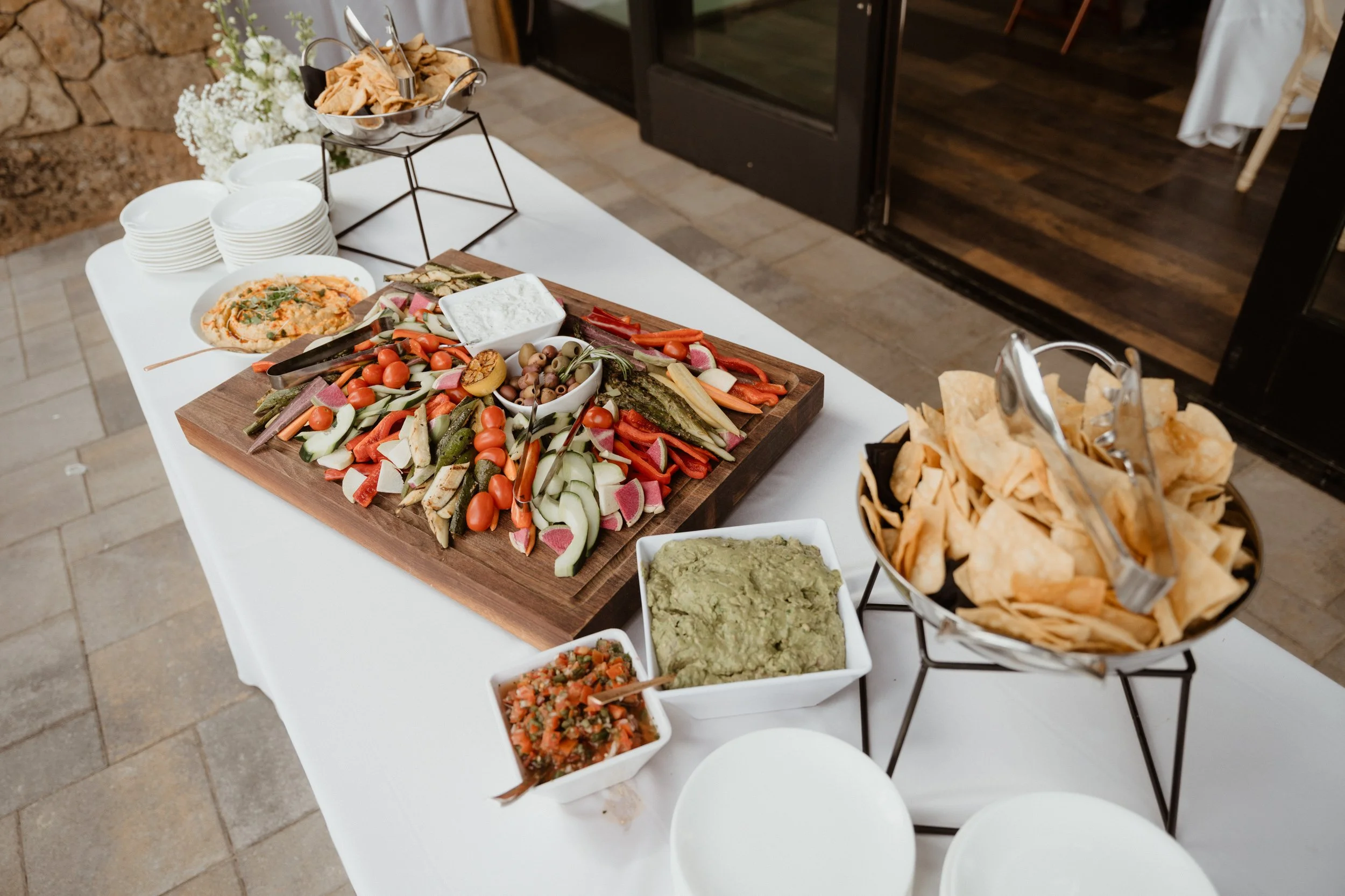 A table of snacks including a large vegetable platter, bowls of guacamole and salsa, a dish of tortilla chips, and a small bowl of hummus, set near a restaurant entrance.