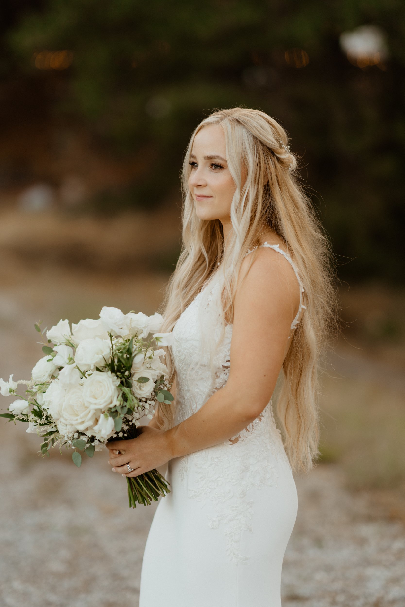 A bride in a white wedding dress holds a bouquet of white roses and greenery, standing outdoors with a blurred natural background.
