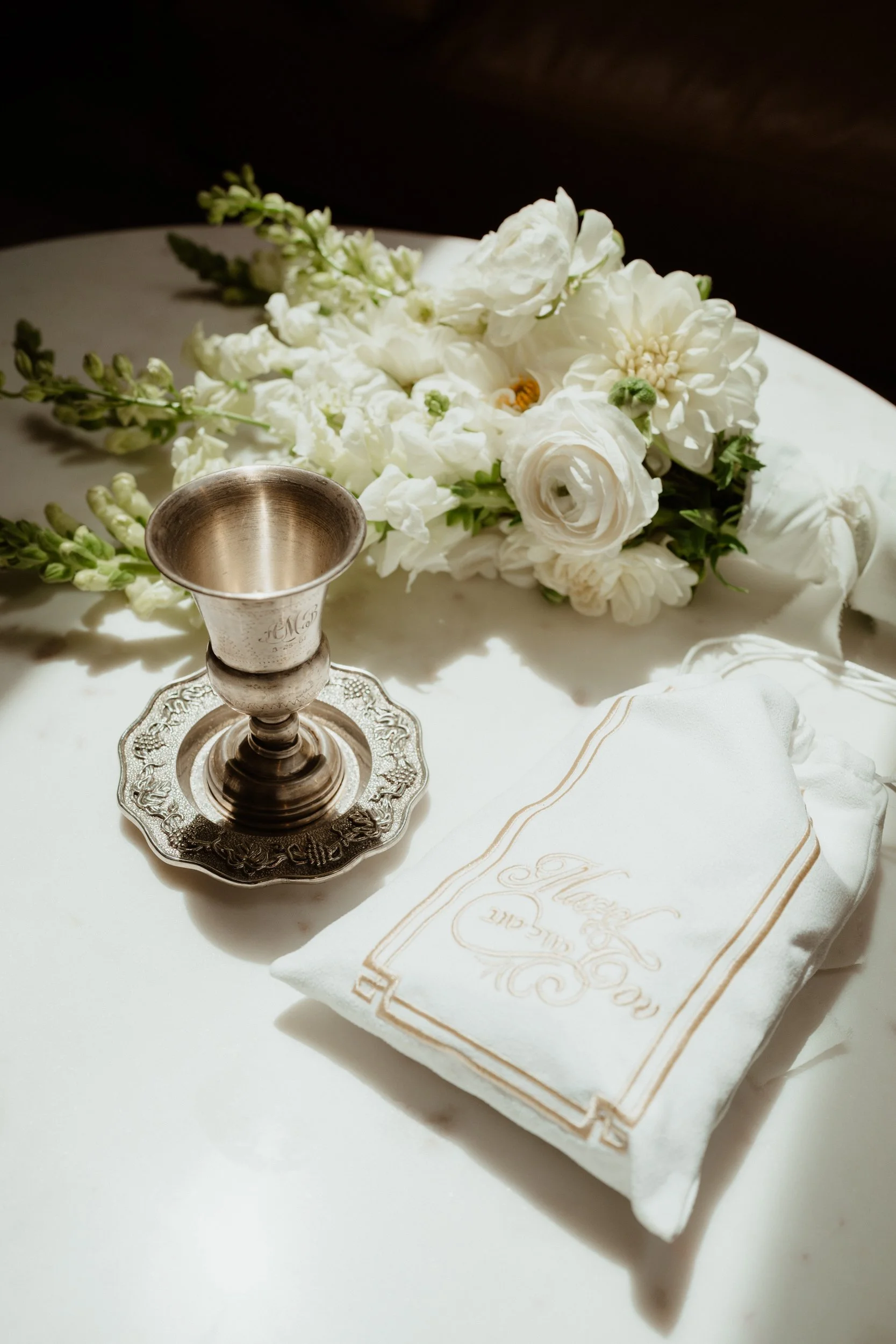 Silver goblet on a decorative silver plate, a white embroidered napkin, and a bouquet of white flowers on a white surface, with soft lighting.