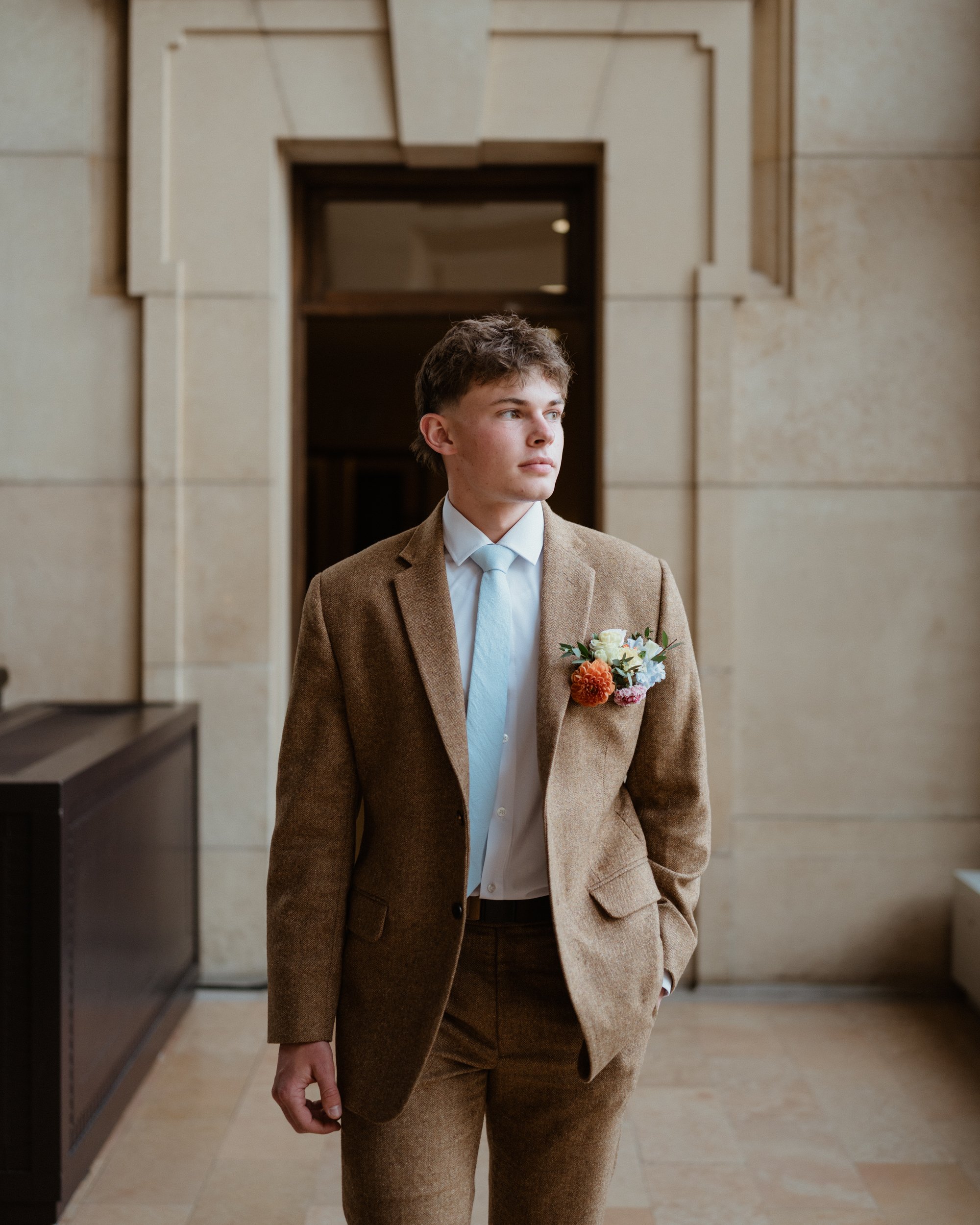 A young man in a brown suit with a light blue tie, standing indoors in front of a wooden door, with a flower boutonniere on his lapel, looking to the side.