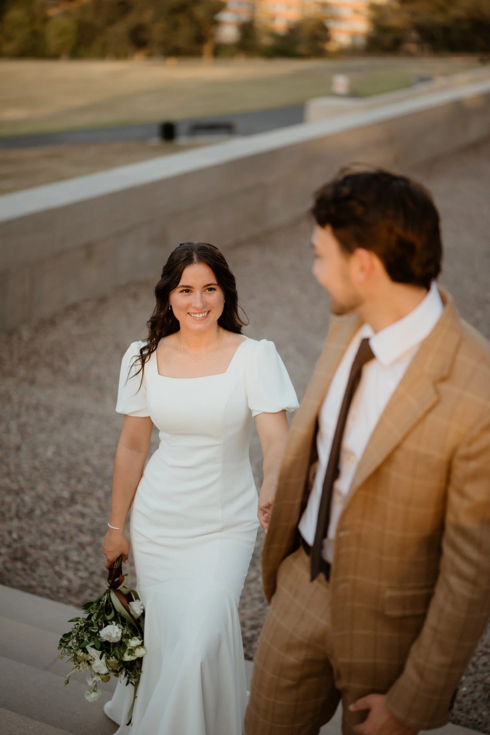 A bride in a white wedding dress holding a bouquet, smiling at a groom in a beige plaid suit, outdoors near steps.