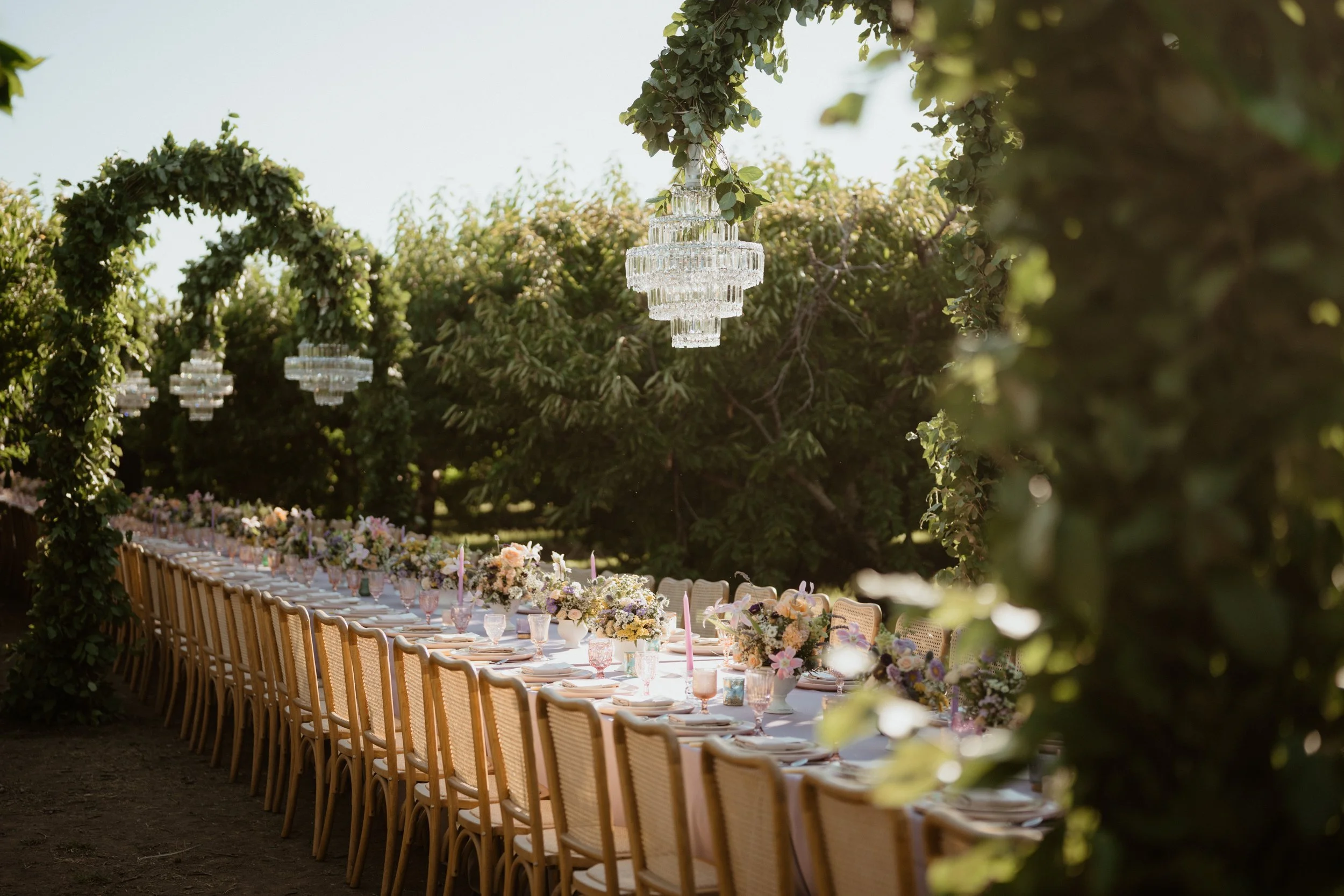 Long outdoor dining table decorated with floral centerpieces and pink glassware, set beneath hanging crystal chandeliers, surrounded by greenery.