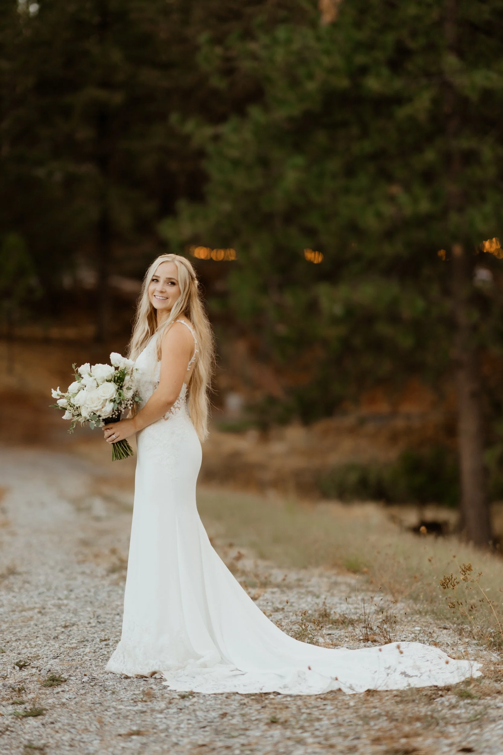A bride in a white wedding dress holding a bouquet of white flowers standing outdoors on a gravel path with trees in the background.