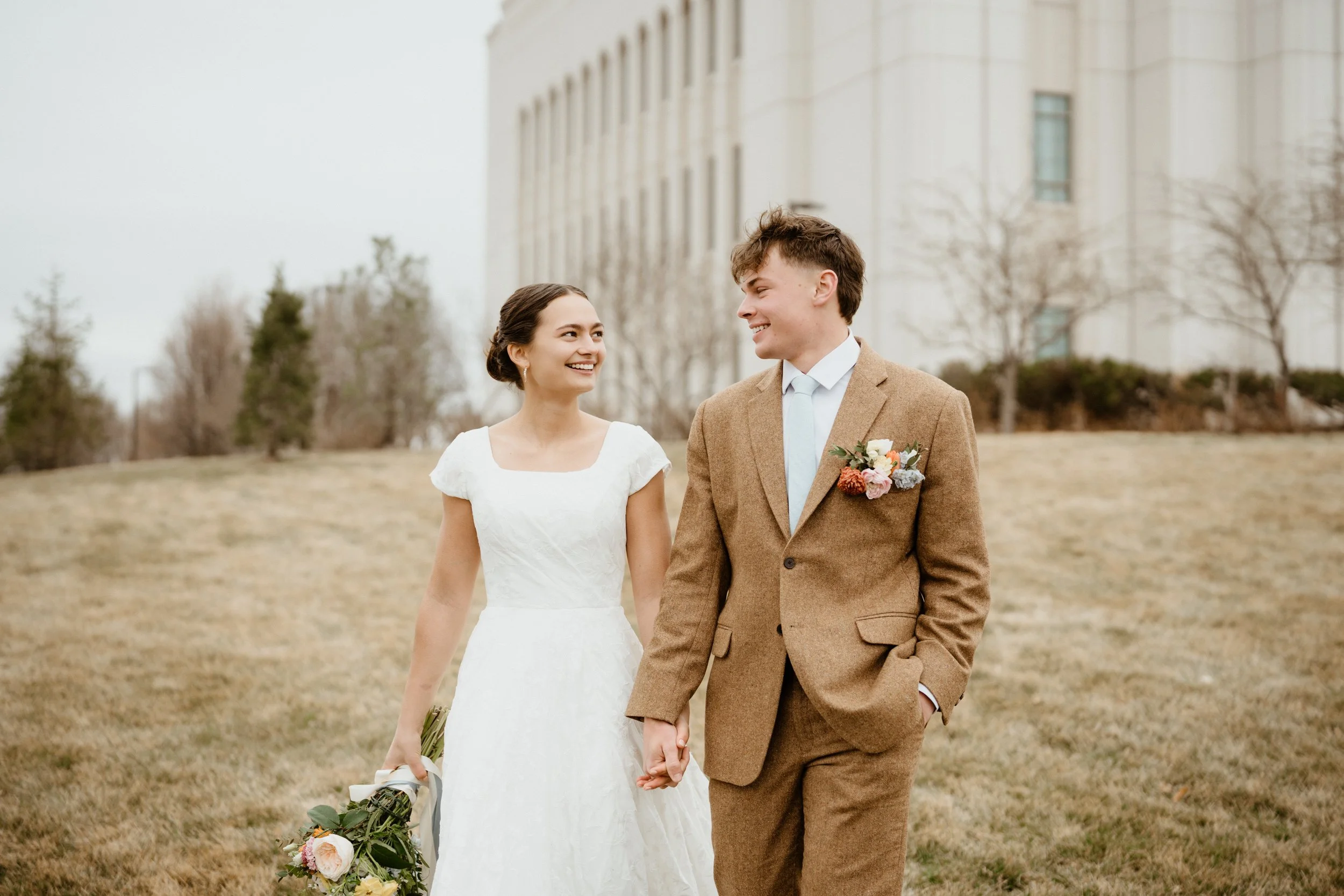A bride and groom walking hand in hand outdoors, smiling at each other, with the bride holding a bouquet and the groom wearing a boutonniere. They are dressed in wedding attire, with a large building in the background.