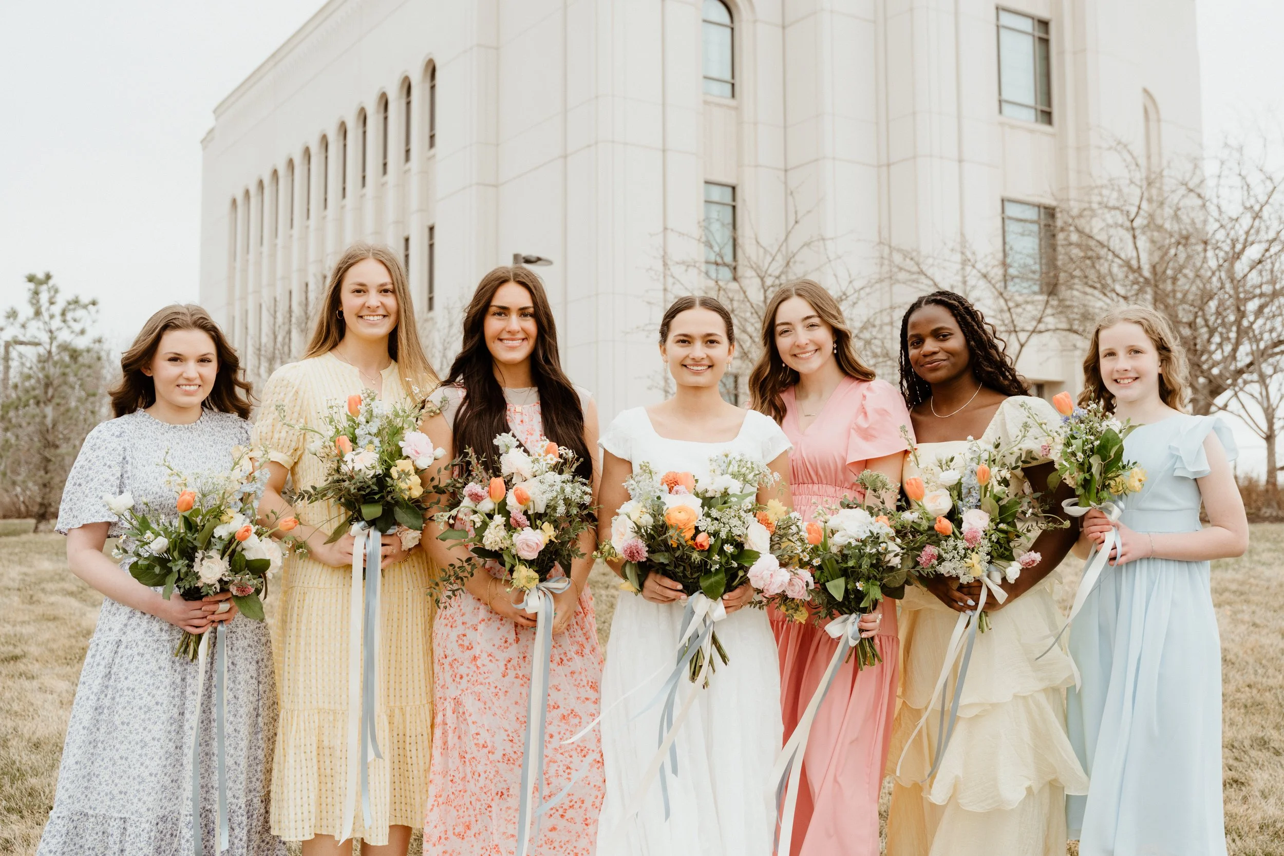 Group of seven young women holding bouquets of flowers outdoors in front of a large building, dressed in pastel and floral dresses, smiling at the camera.