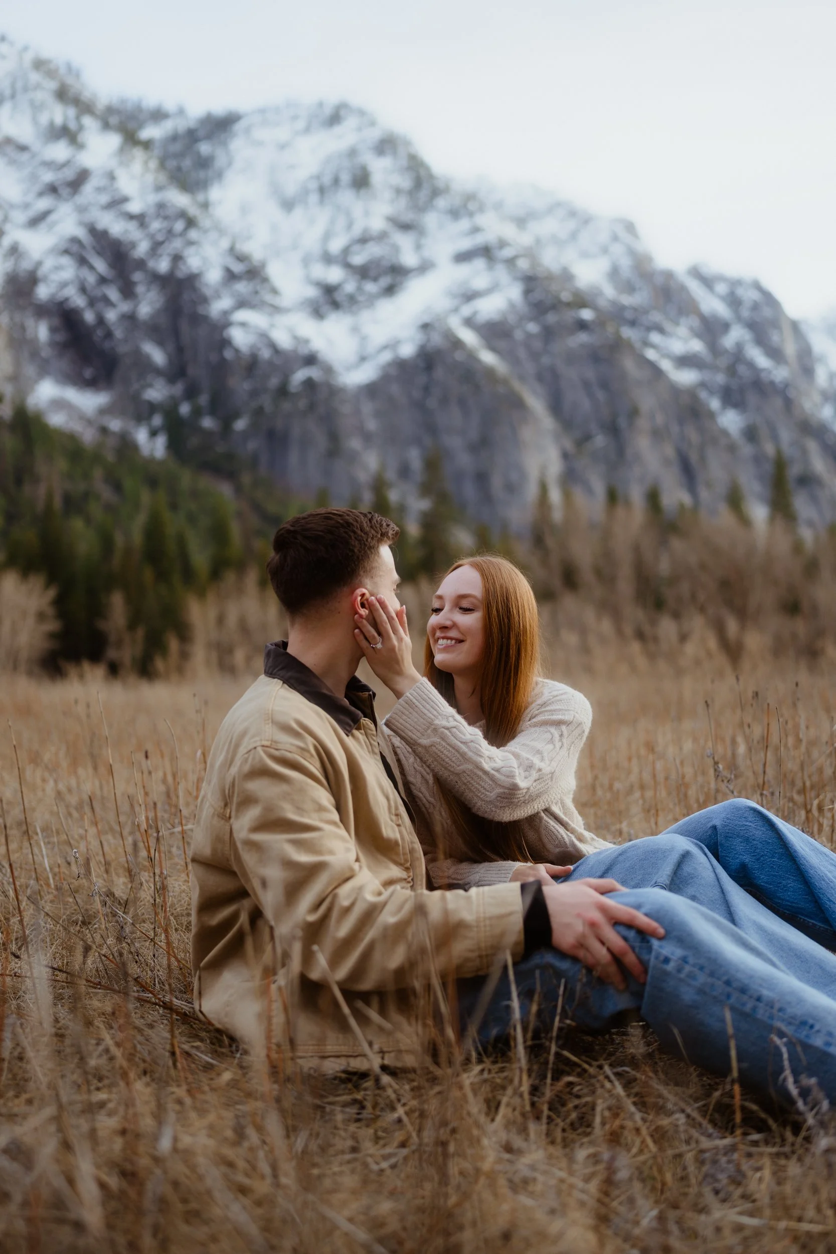 A couple sitting in a field with mountains and snow in the background, smiling and interacting with each other.