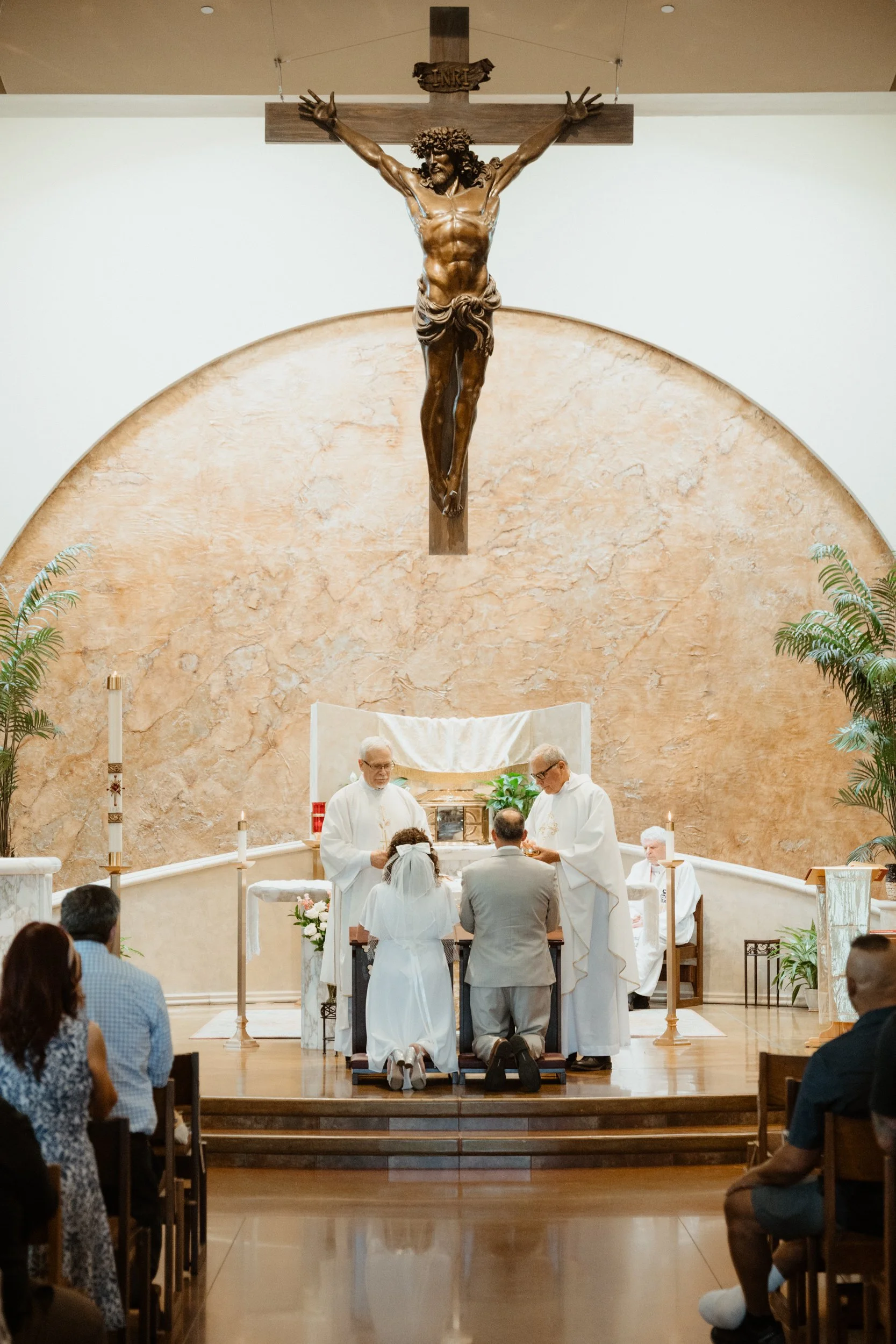 A wedding ceremony in a church with a priest and a couple kneeling at the altar, with a large crucifix of Jesus Christ hanging above.
