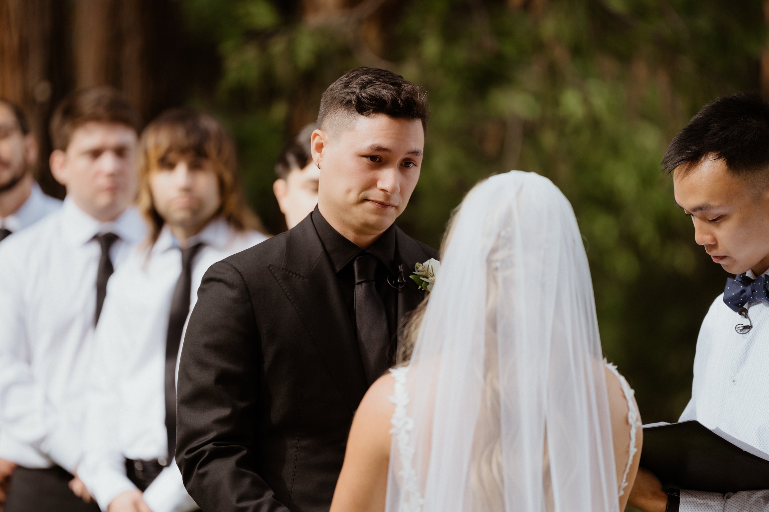 Man in a black suit and tie looking at a woman in a wedding dress and veil, standing outdoors, with several men in white shirts and black ties in the background.
