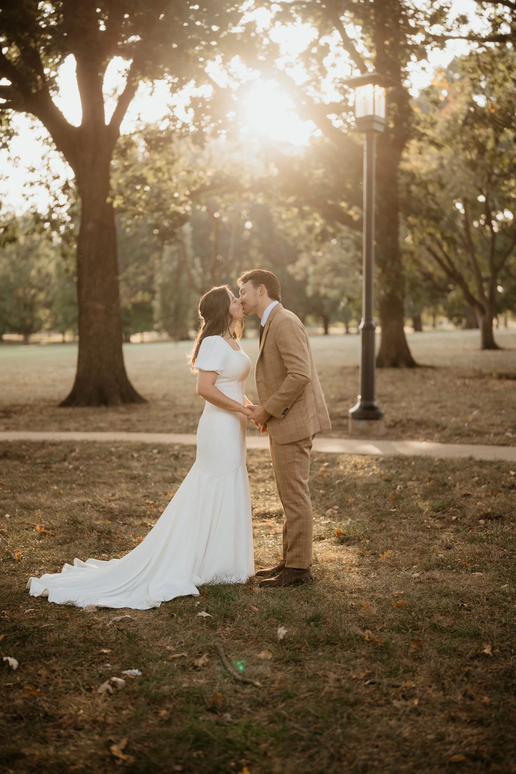 A newlywed couple sharing a kiss in a park at sunset, holding hands, with trees and a lamppost in the background.
