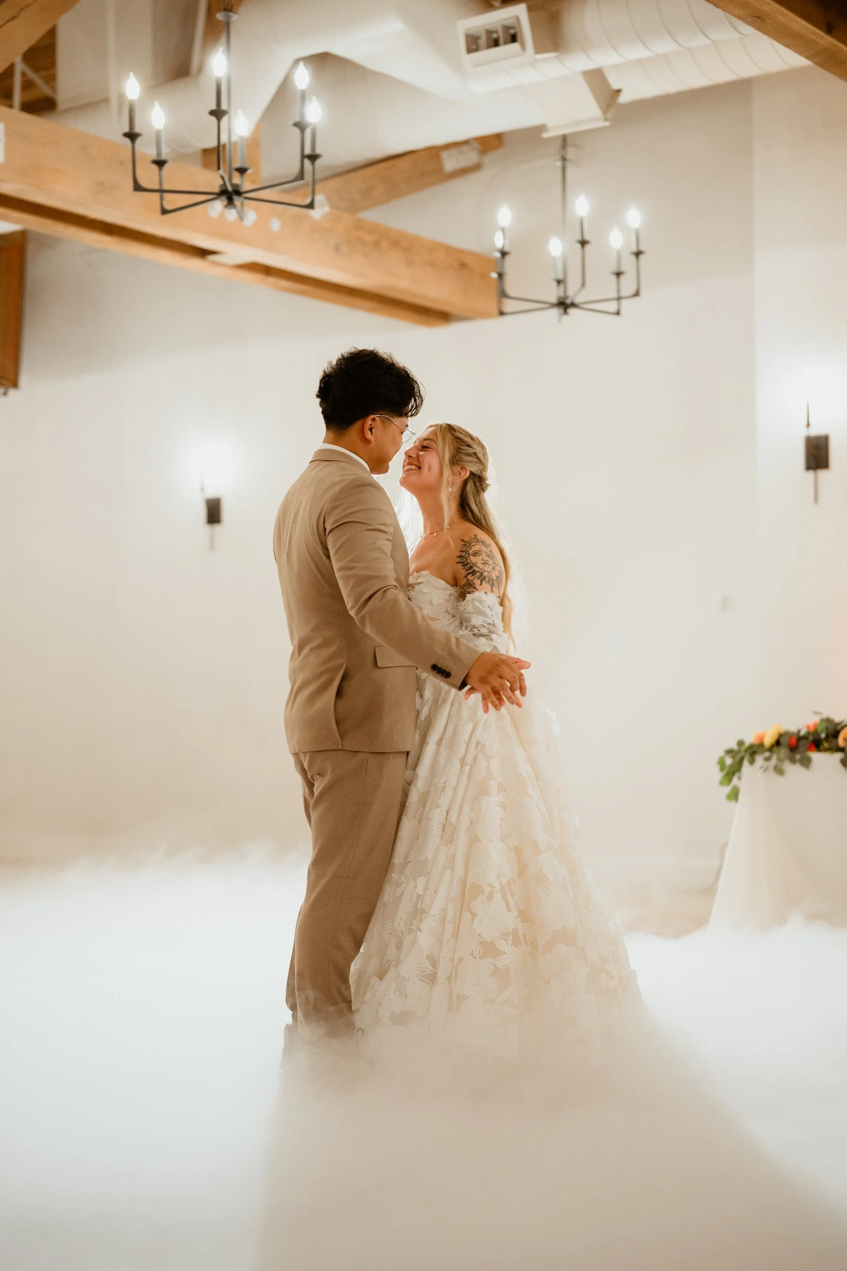 A newlywed couple on the dance floor, holding hands and smiling at each other, during their wedding reception in a warmly lit room with wooden beams and chandeliers.