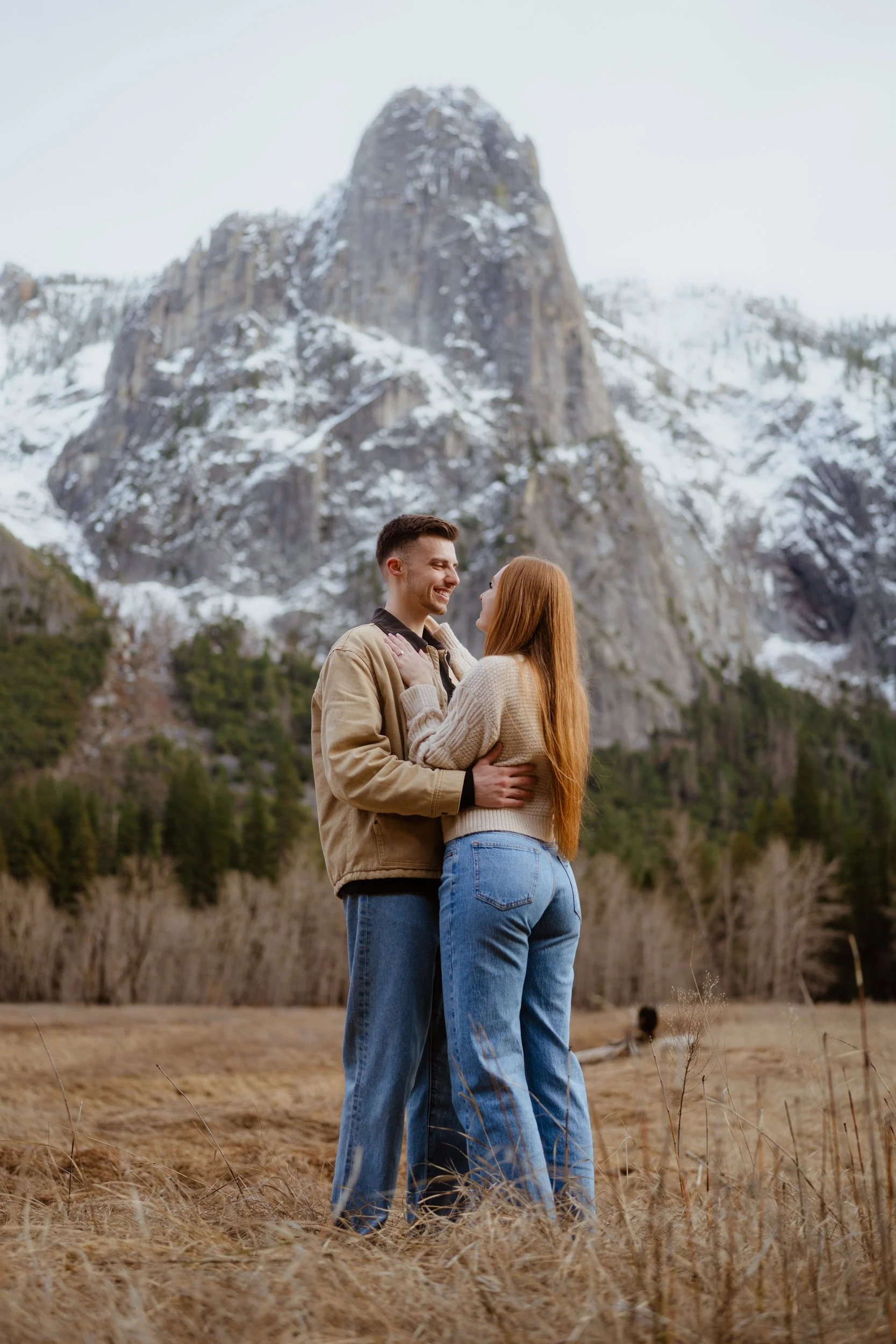 A young couple stands close together in a grassy field, smiling at each other, with a large snow-capped mountain in the background.