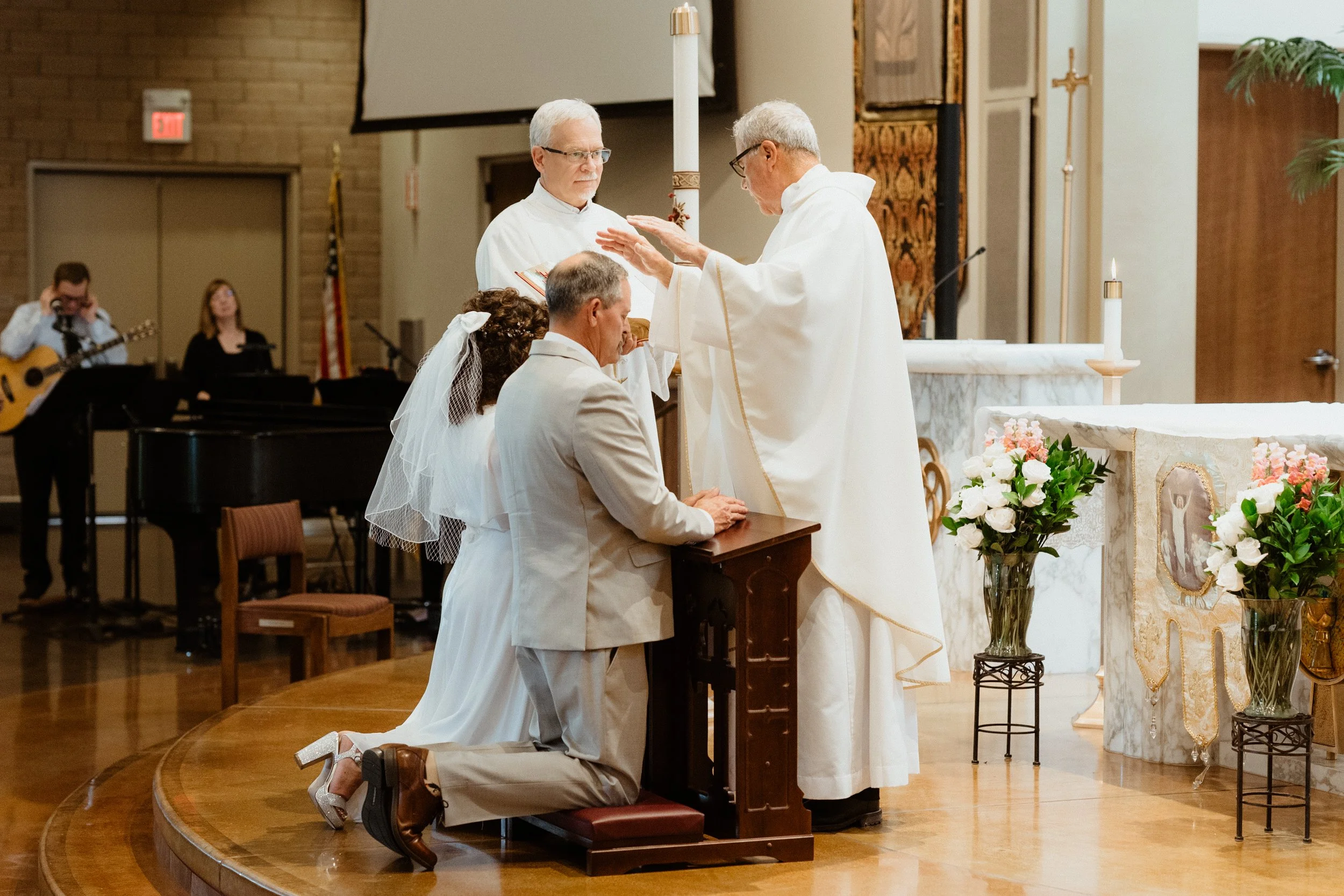 A wedding ceremony with a priest officiating a couple's vows. The kneeling groom is in a beige suit, and the bride is in a white dress with a veil, kneeling on a platform. Two assistants stand behind the bride and groom, with a drummer and guitarist 