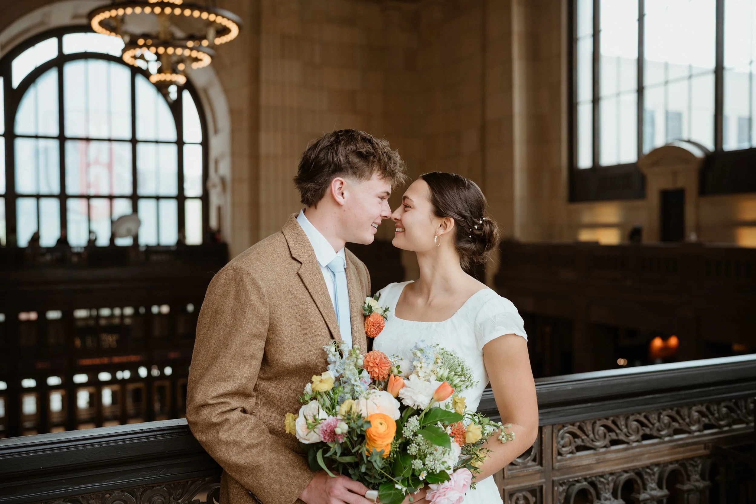 A bride and groom standing close together inside a church, smiling and touching noses. The bride holds a colorful bouquet of flowers, and the groom is wearing a brown suit and light blue tie.