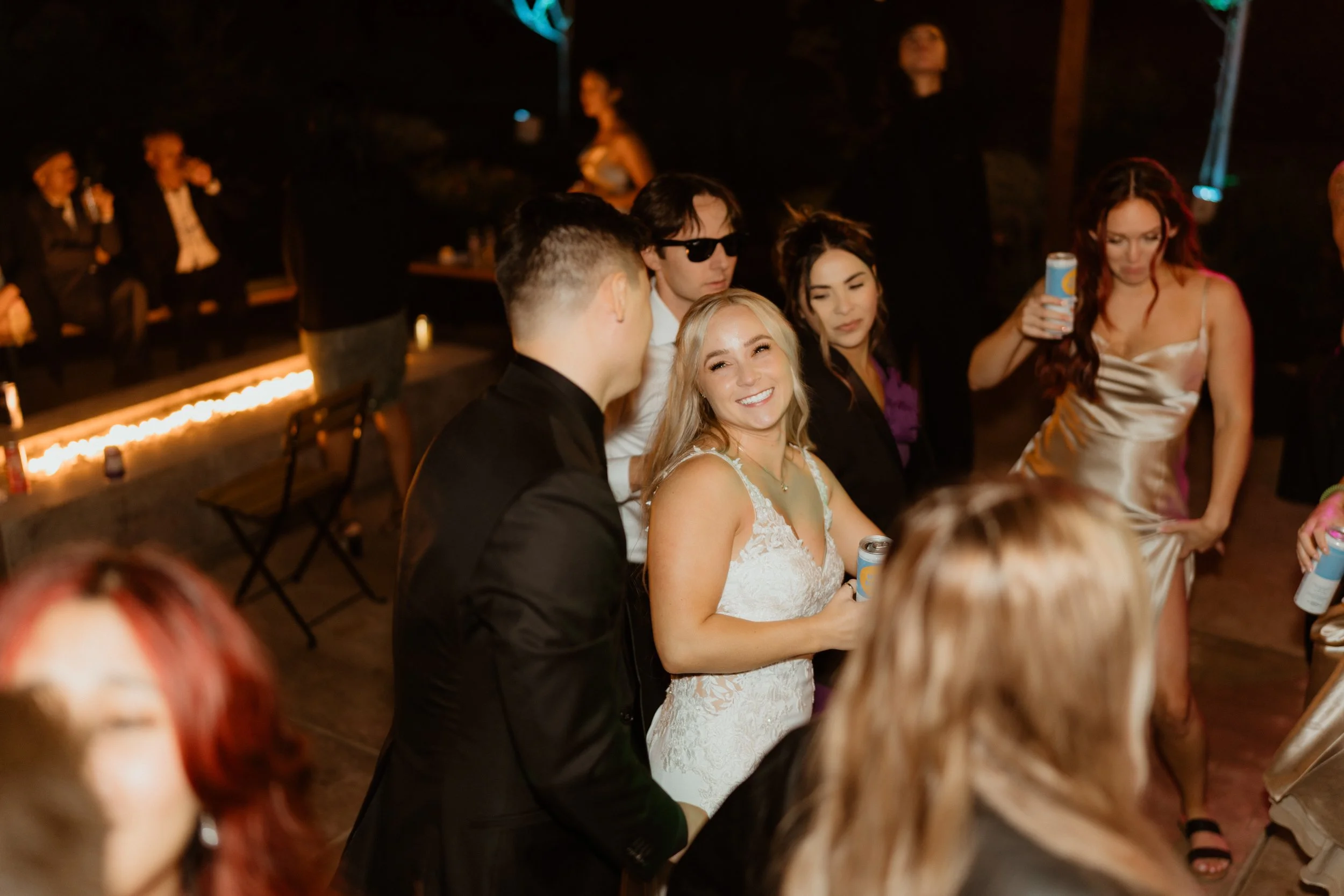 People dancing and socializing at a nighttime wedding reception, including a smiling bride in a white wedding dress holding a drink, surrounded by friends in formal attire.