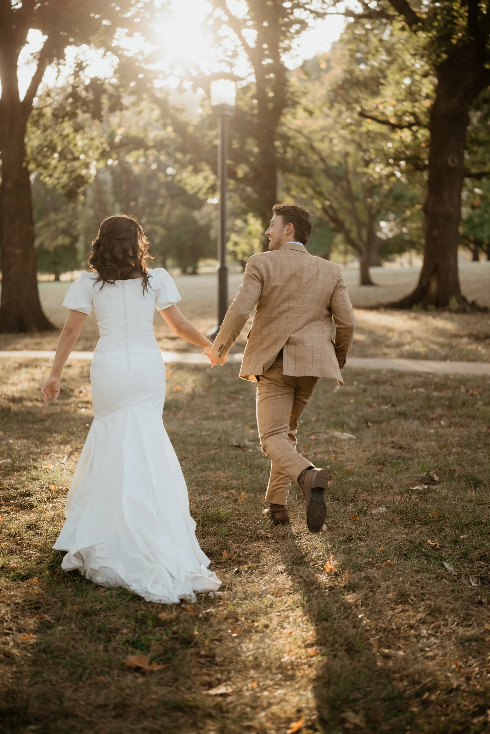 A newlywed couple holding hands and running through a park with trees and sunlight in the background.