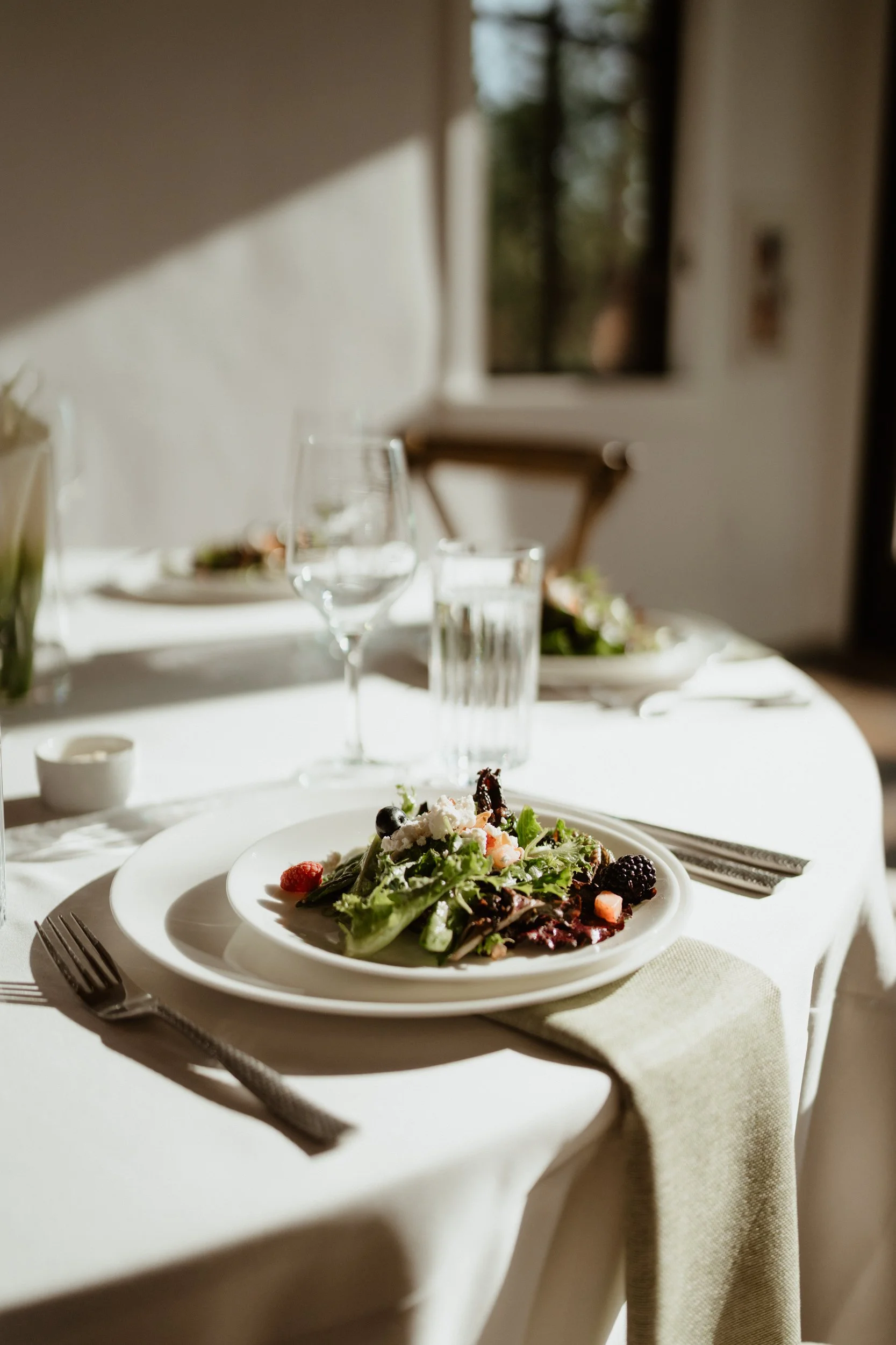 A table with a white tablecloth set for a meal, featuring a salad with berries and cheese on a plate, a fork, and a napkin in a well-lit room with natural sunlight.