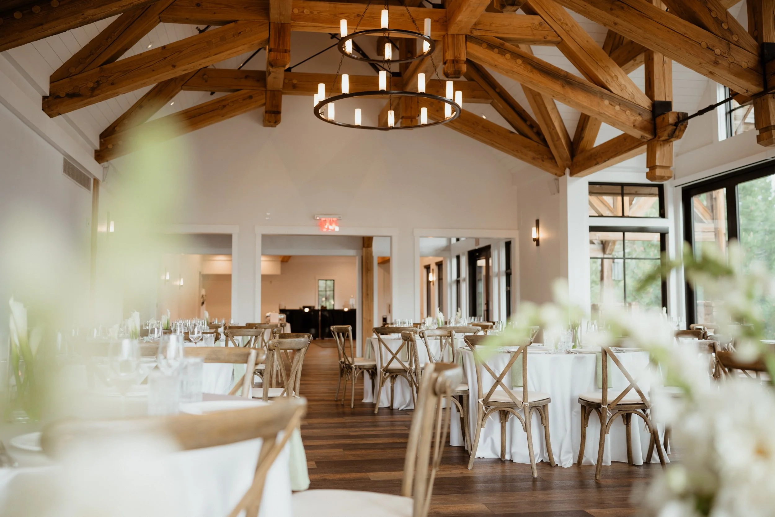Elegant event venue with wooden beams, large windows, round tables with white tablecloths, and wooden chairs, decorated for a celebration.