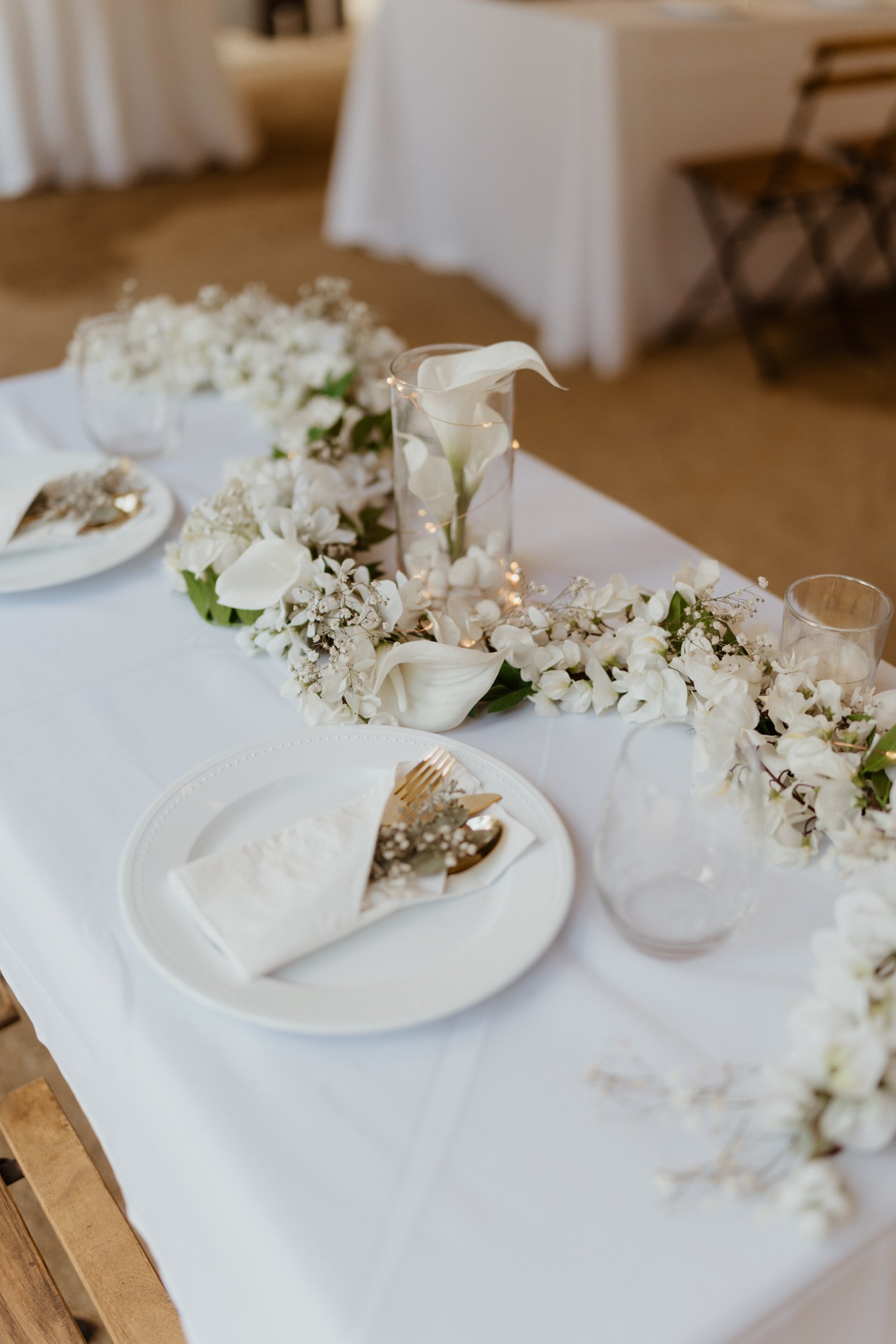 A decorated dining table with white flowers, a tall glass candle holder with a white calla lily, plates with white napkins, and clear glass cups.