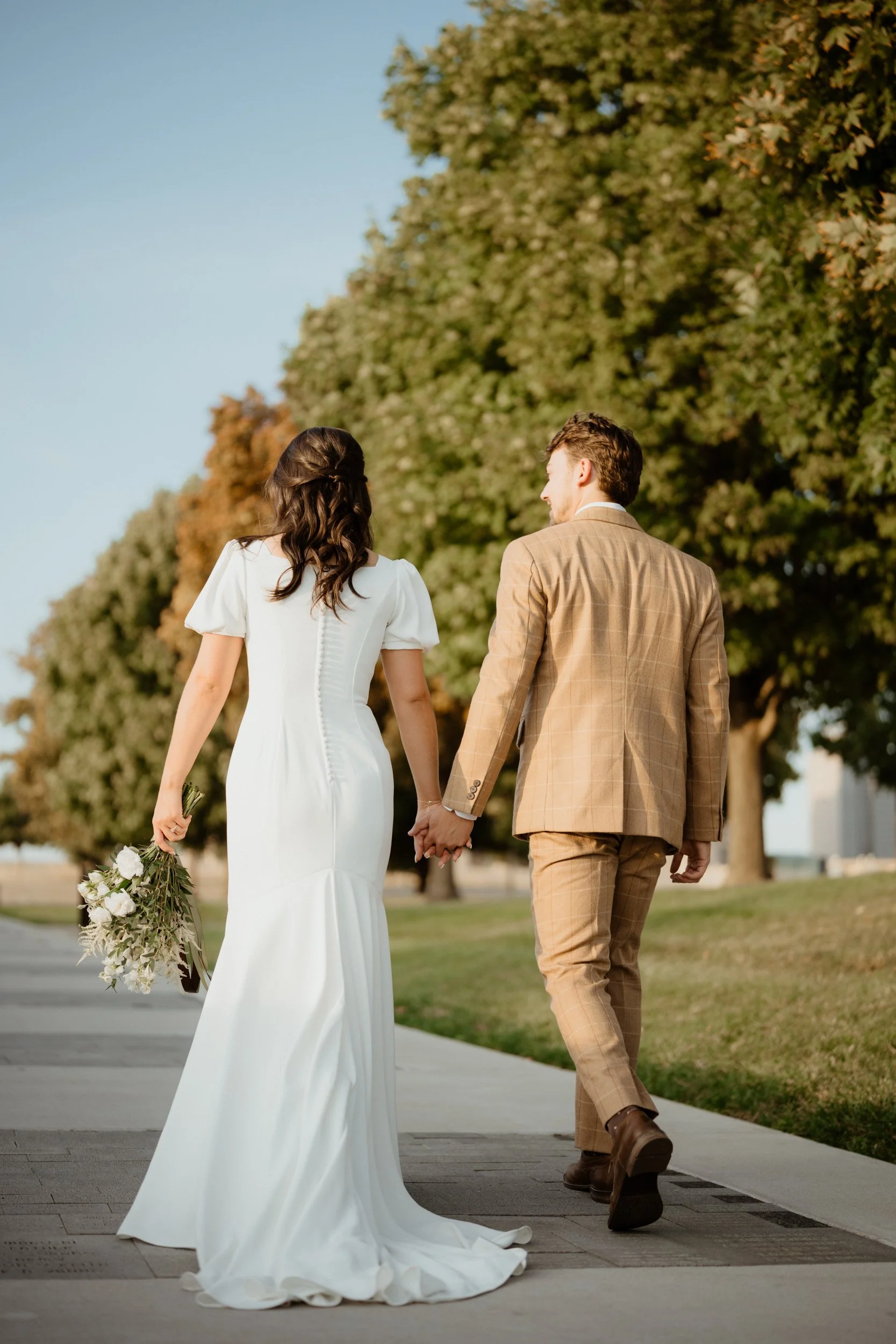 A bride and groom holding hands while walking outdoors on a sunny day, with trees in the background. The bride is wearing a white wedding dress and carrying a bouquet, and the groom is dressed in a beige plaid suit.