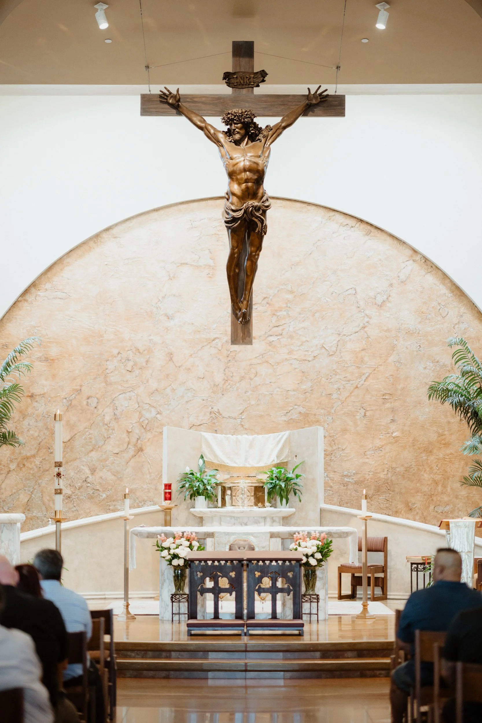 Interior of a chapel with a large crucifix of Jesus Christ hanging above the altar, which is decorated with flowers and plants, and surrounded by candles and chairs.