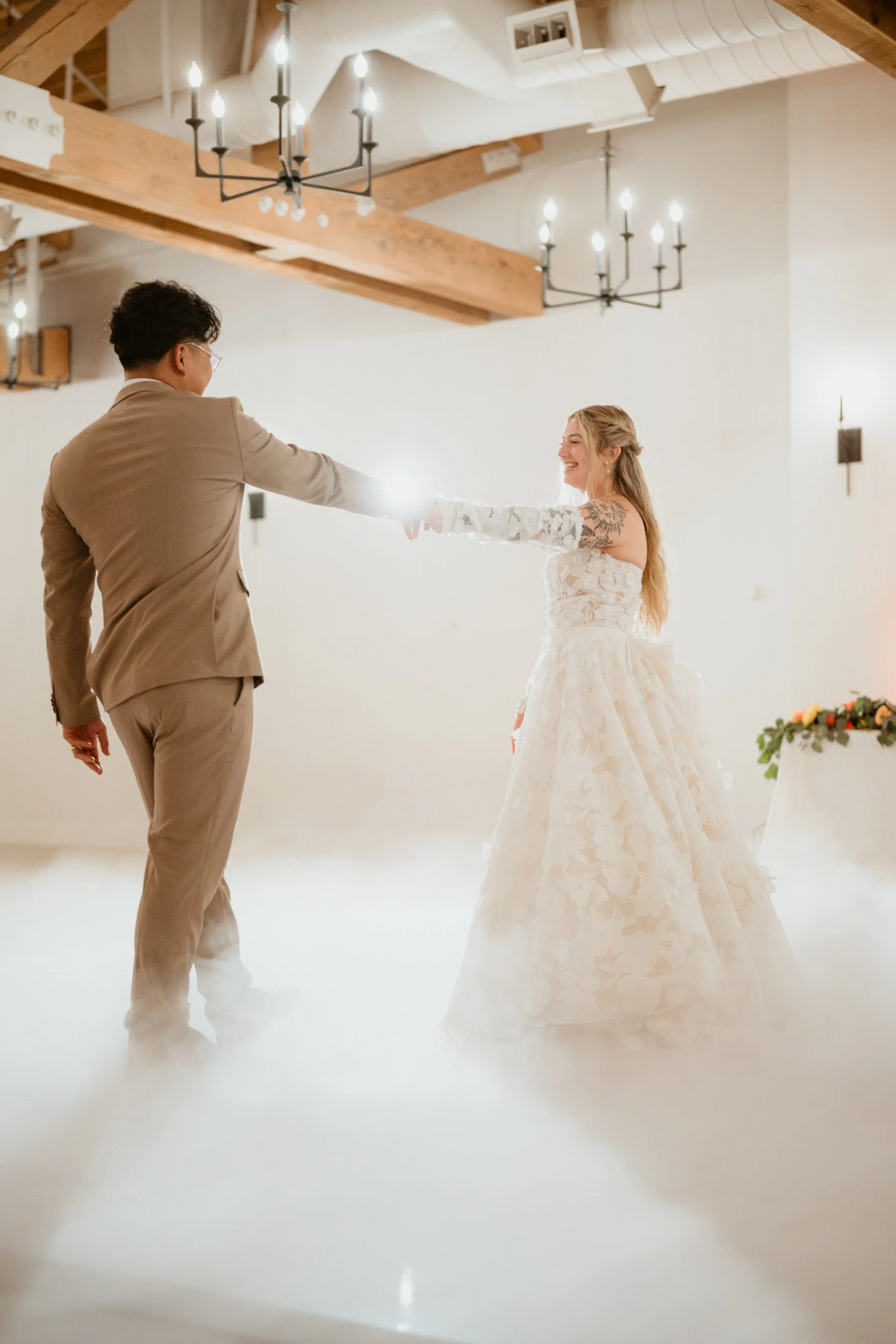 A bride and groom dancing at their wedding reception in a decorated indoor venue with wooden beams and chandeliers, with fog effects on the floor.