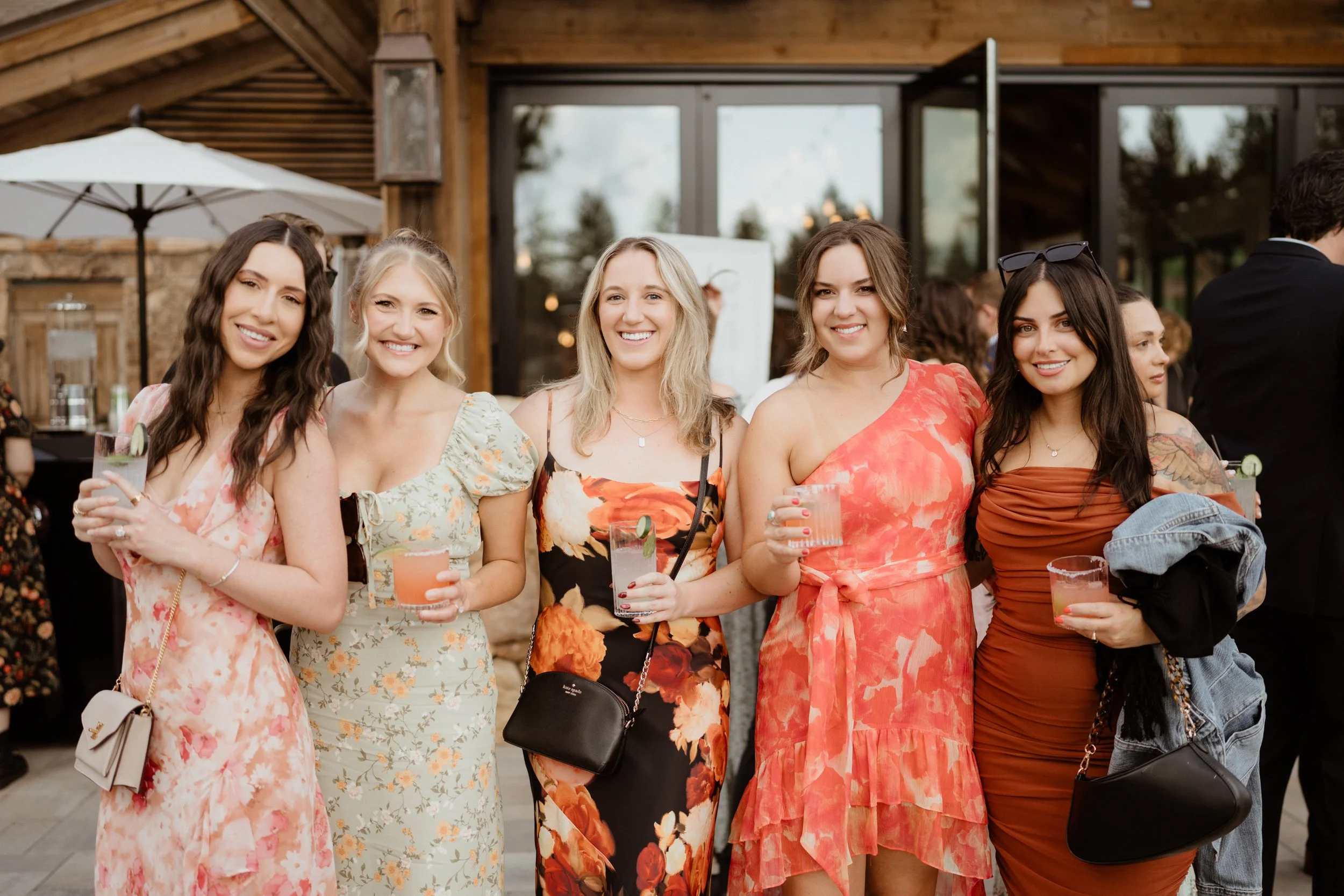 Five women standing together at an outdoor event, smiling and holding drinks, dressed in colorful floral and solid-colored dresses.