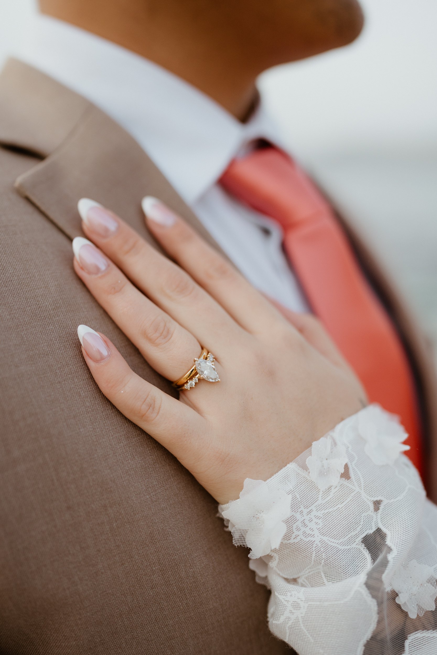 Close-up of a woman's hand with a wedding ring resting on a man's shoulder. The ring features a large pear-shaped diamond, and the woman is wearing a lace sleeve. The man is wearing a suit with a white shirt and a red tie.