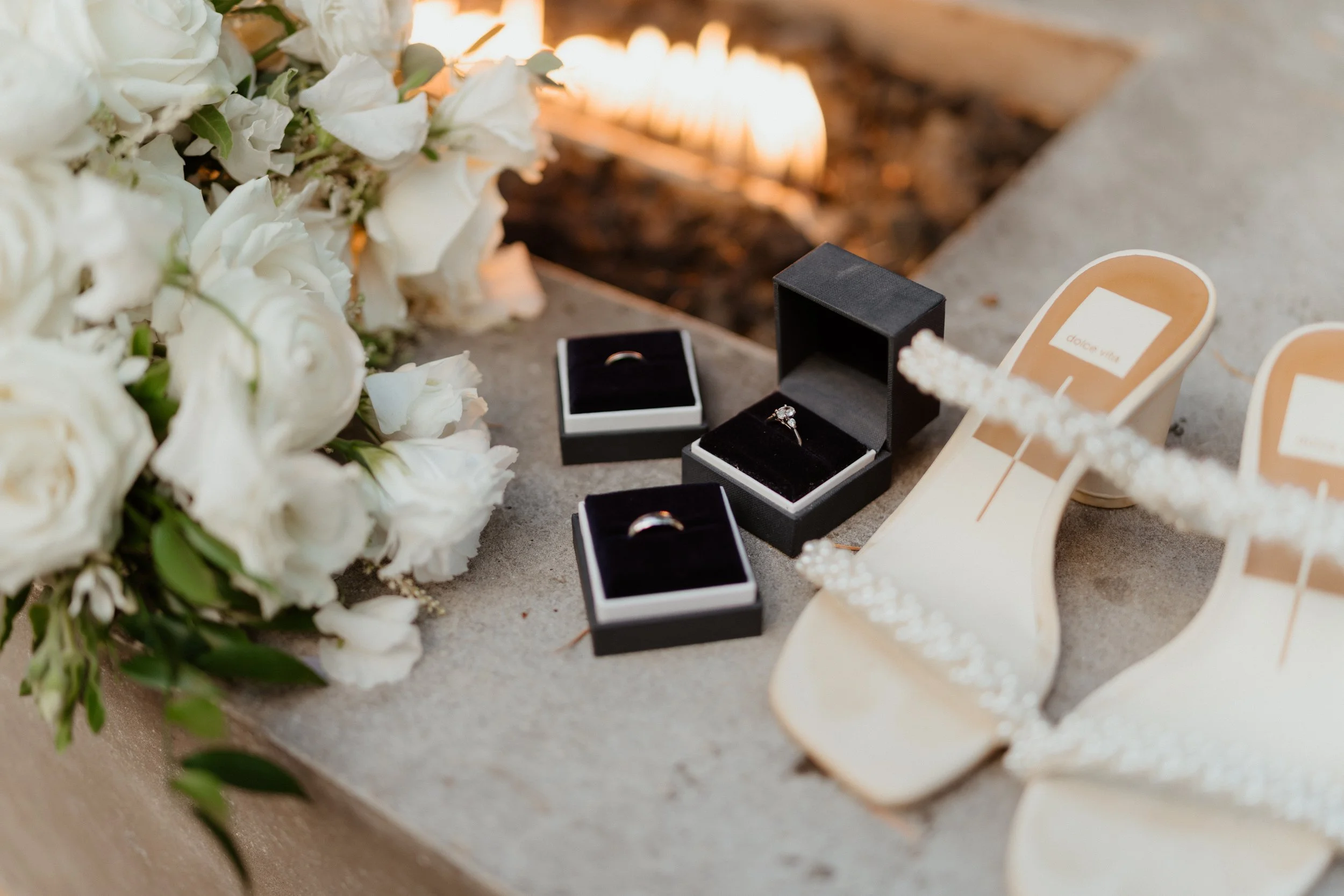 Wedding rings displayed in black and beige boxes, white high-heeled shoes with pearl details, and a bouquet of white flowers on a concrete surface.