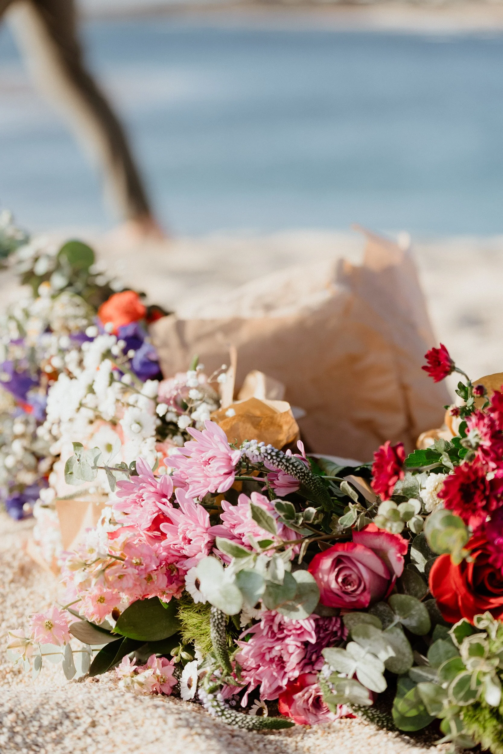 Colorful bouquet of flowers, including roses and daisies, resting on sandy beach with ocean in background.