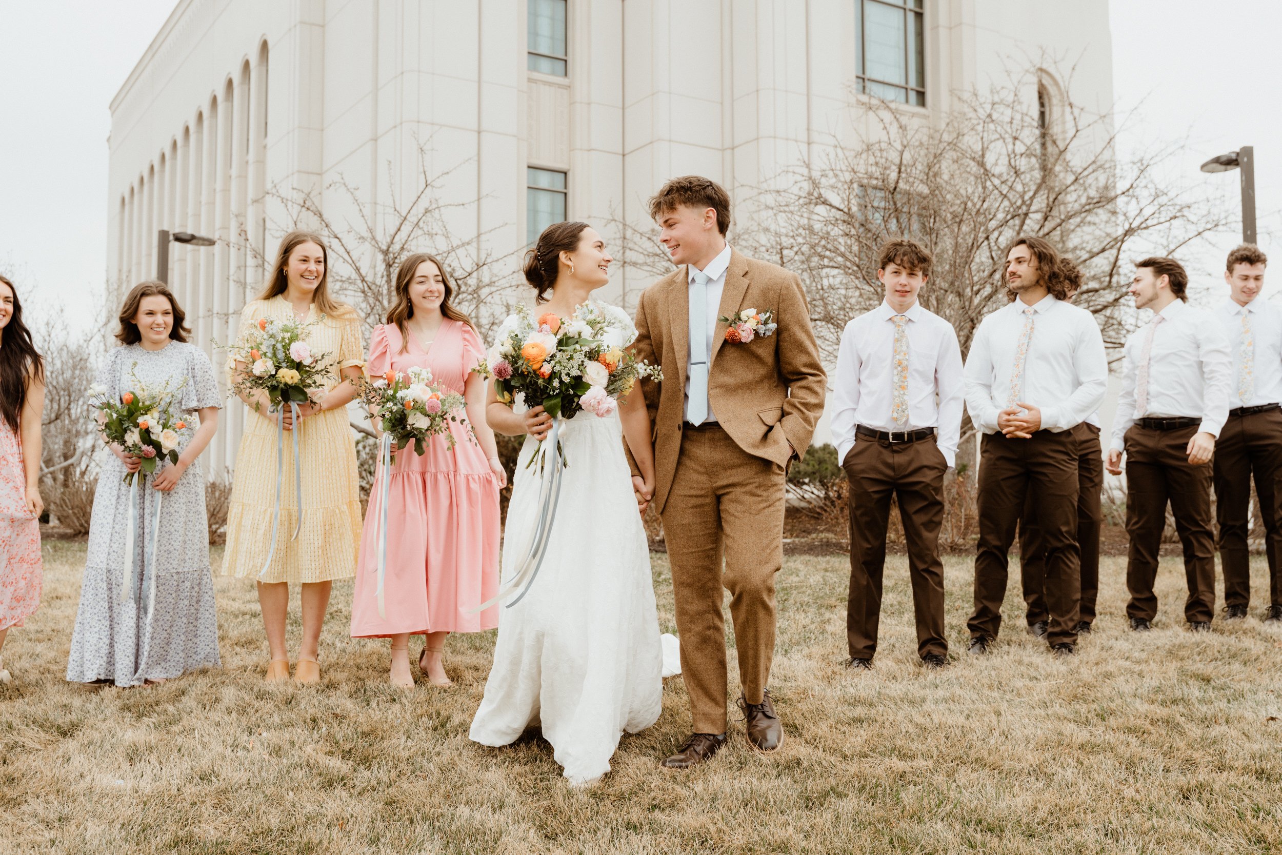A wedding ceremony outdoors with a bride and groom holding hands, surrounded by bridesmaids and groomsmen standing on grass, with a large building and leafless trees in the background.