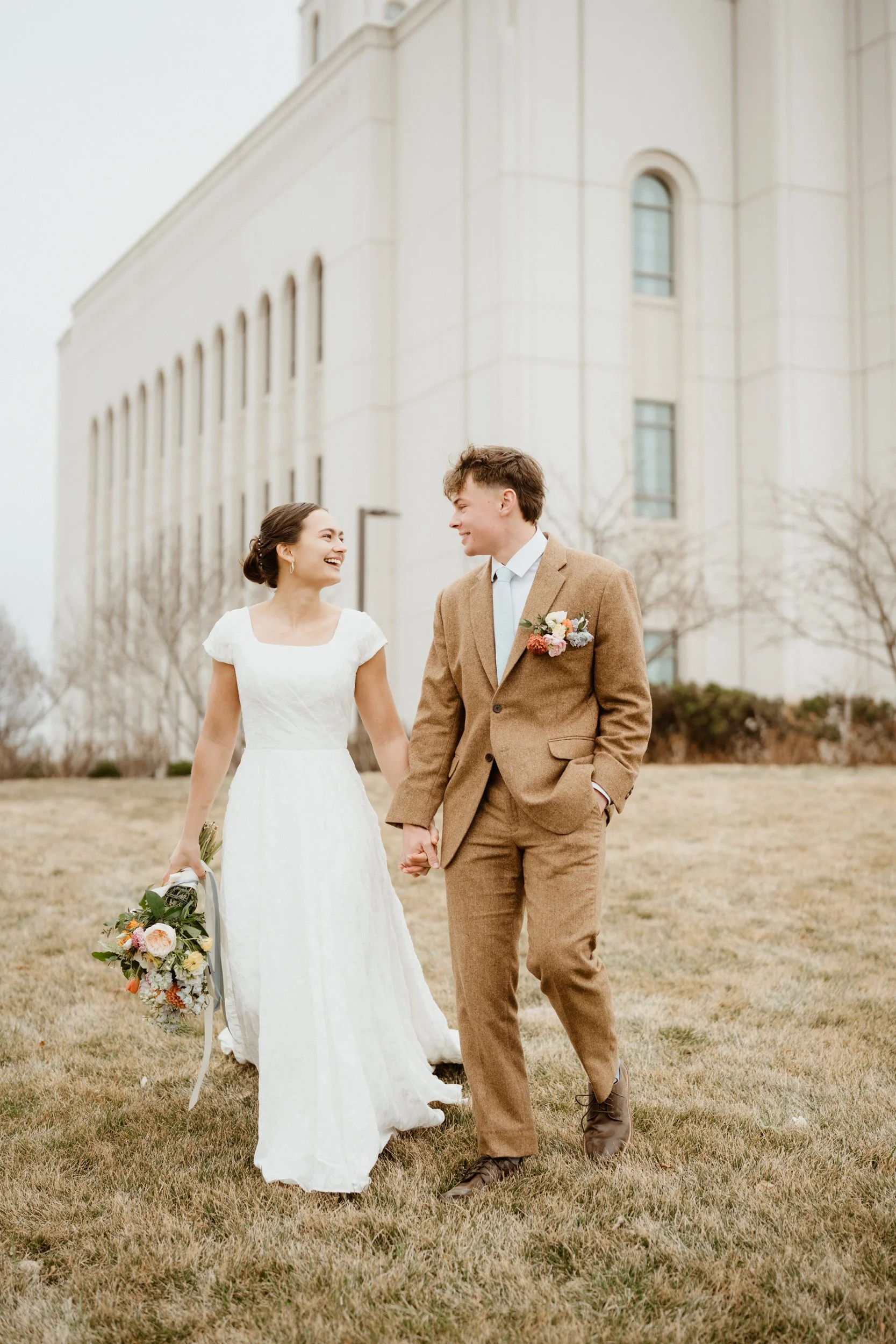 A happy bride and groom holding hands, walking on grass in front of a large building, smiling at each other.
