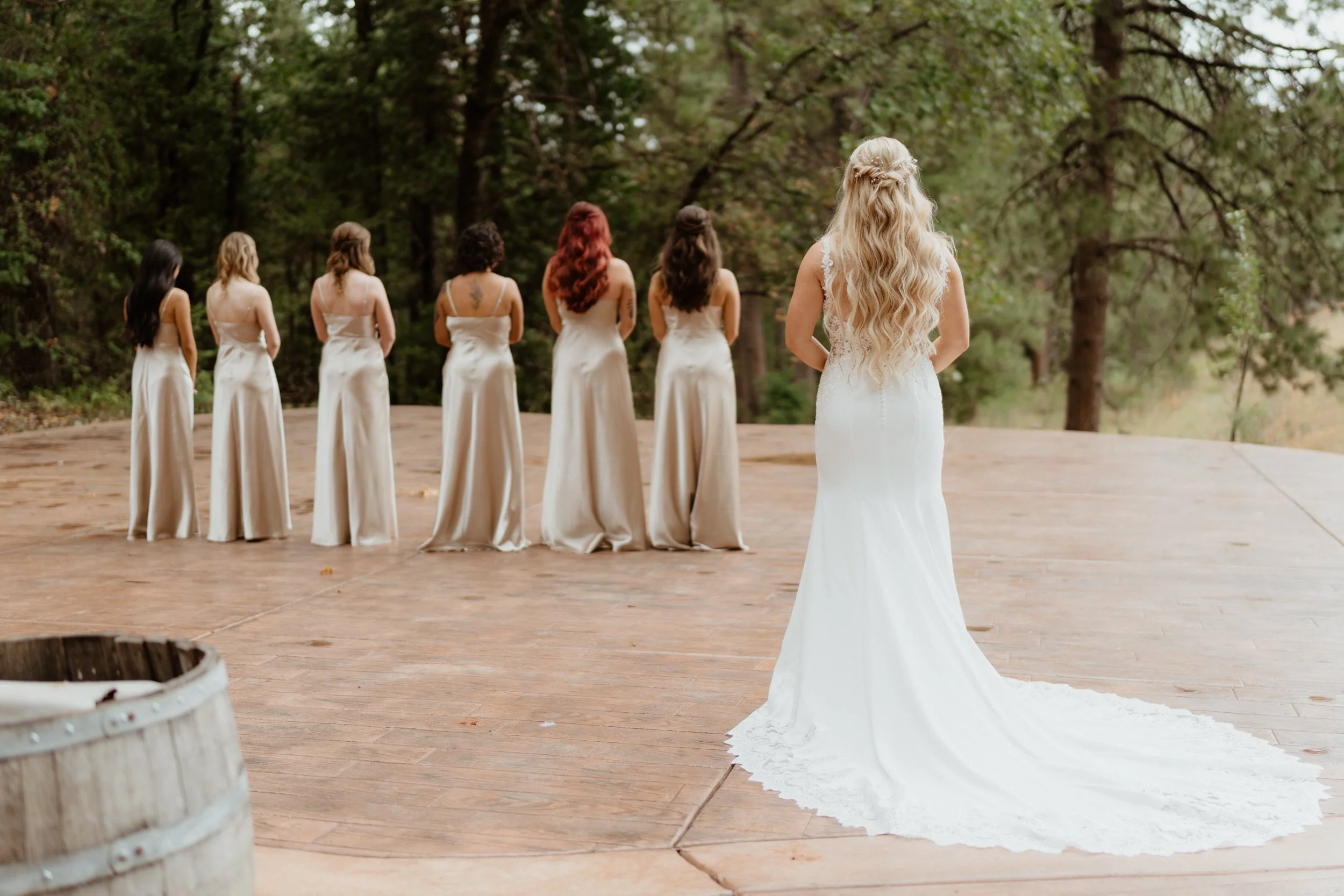 Bride in a white wedding dress watching her bridesmaids, who are lined up in white dresses, outside in a wooded area on a wooden platform.