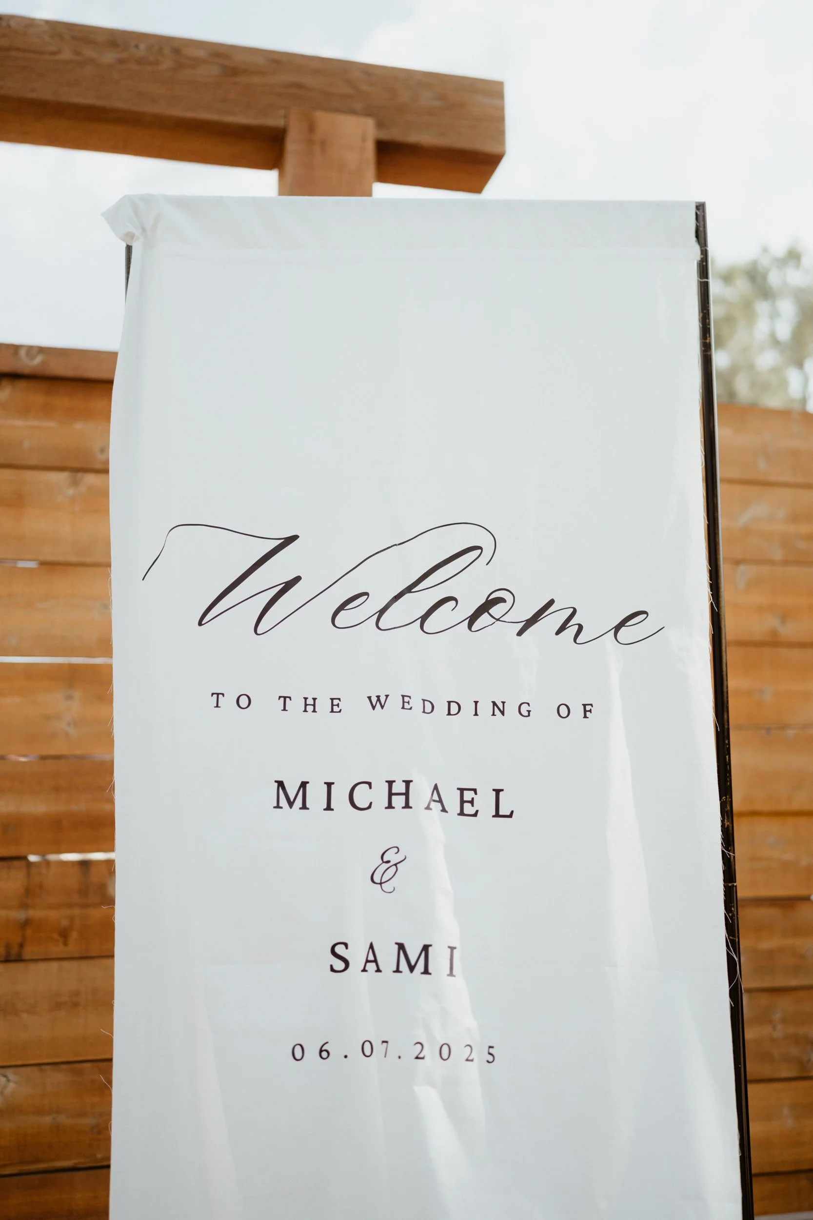 Wedding welcome sign for Michael and Sami, dated 06.07.2025, with script and serif font on a white background, against a wooden fence.