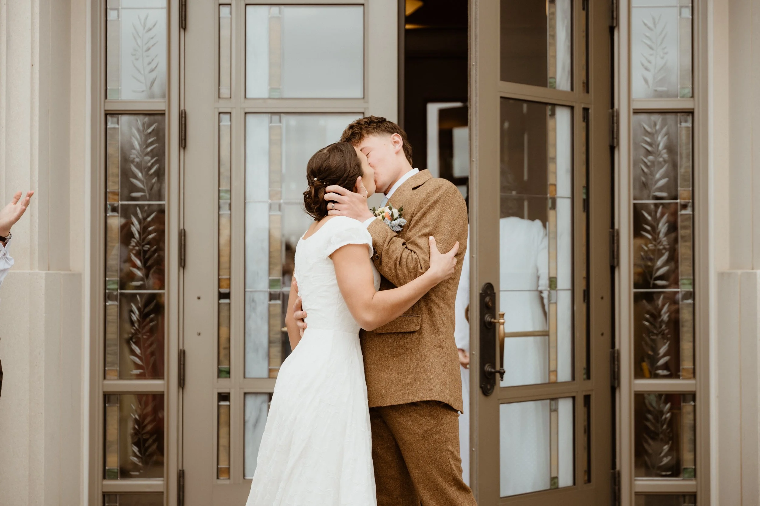 A couple in wedding attire sharing a kiss outside a building with glass-paneled doors.